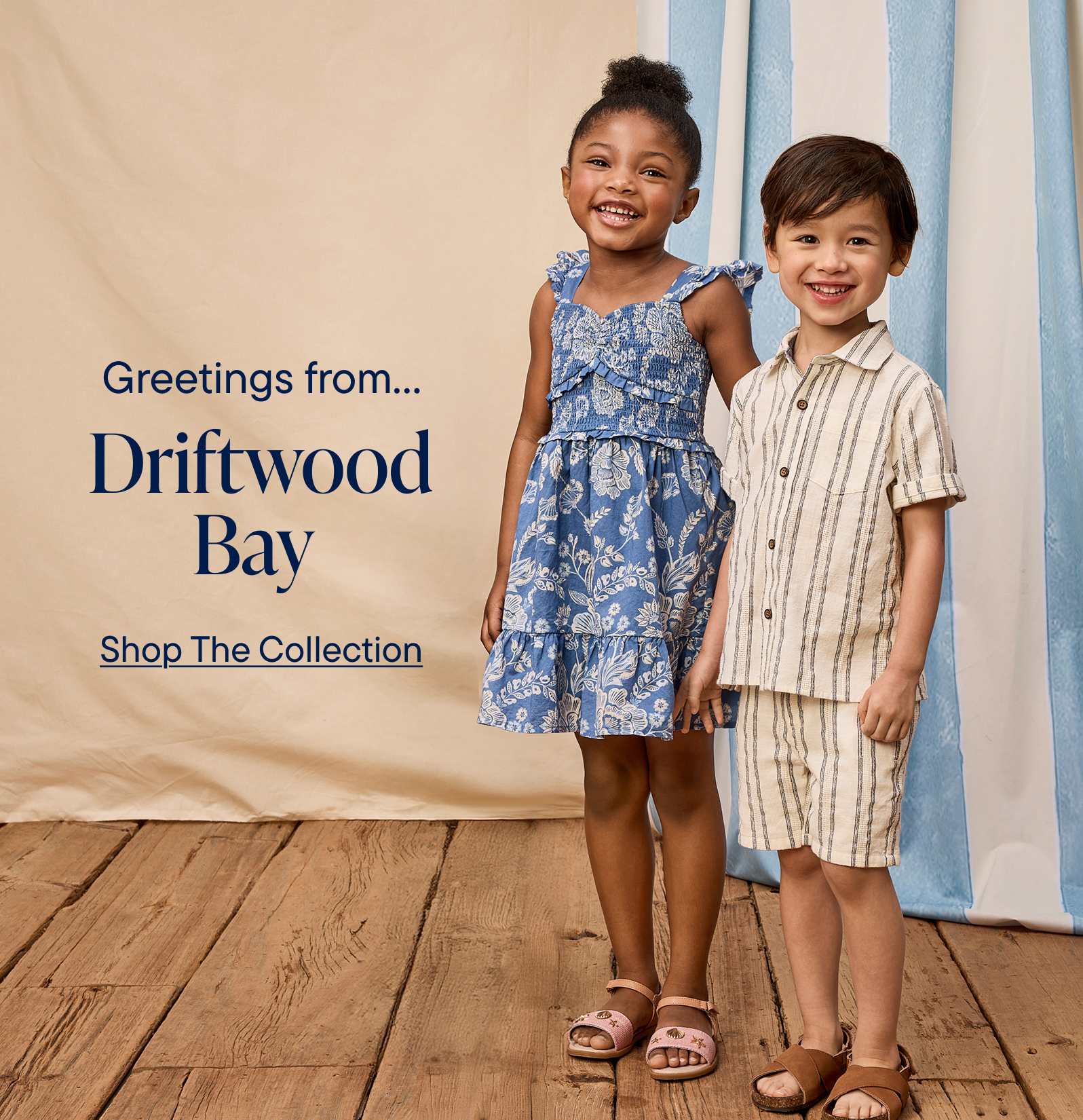 Two smiling children, a girl in a floral dress and a boy in a striped shirt, stand in front of a banner advertising "Greetings from Driftwood Bay" and promoting a clothing collection.