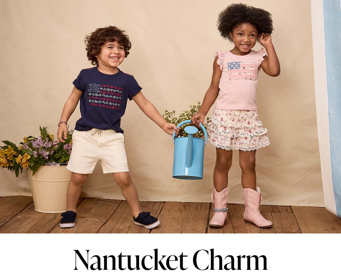 Two young girls with curly hair, one wearing a navy blue shirt and the other a pink top, standing in a room with potted plants in the background.