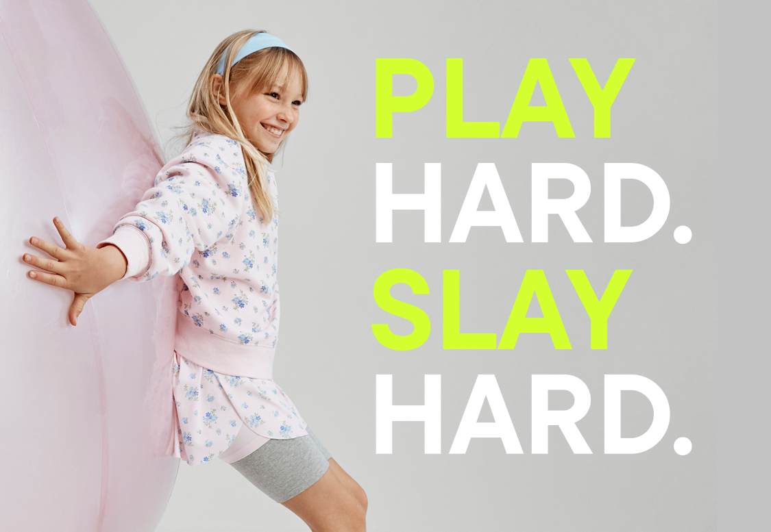 La imagen presenta el texto "PLAY HARD. SLAY HARD" en letras negritas con efecto neón sobre un fondo claro y liso. Se pueden ver unos auriculares en la esquina inferior derecha del marco.