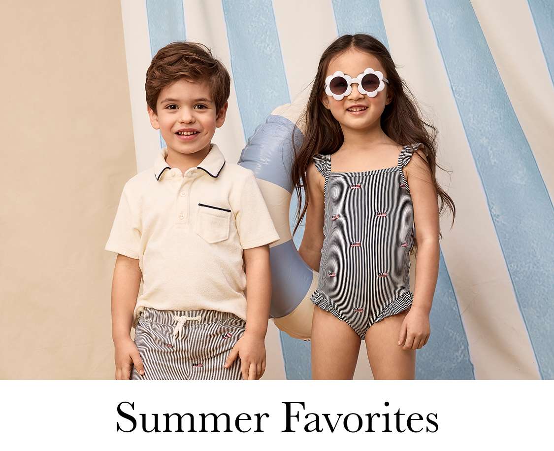 Two young girls with curly hair, one wearing a navy blue shirt and the other a pink top, standing in a room with potted plants in the background.