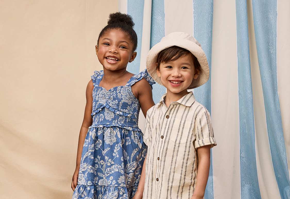 Two young girls, one wearing a white dress and the other a lavender dress, stand on a wooden floor in front of blue and white curtains.