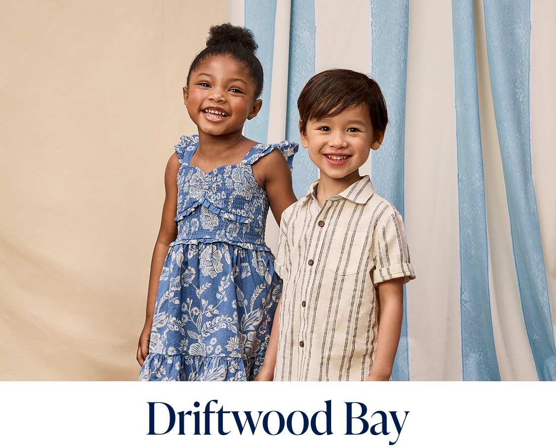 Two young girls, one with long dark hair and the other with short curly hair, are standing on a wooden floor in front of blue and white curtains, with flowers in the background.