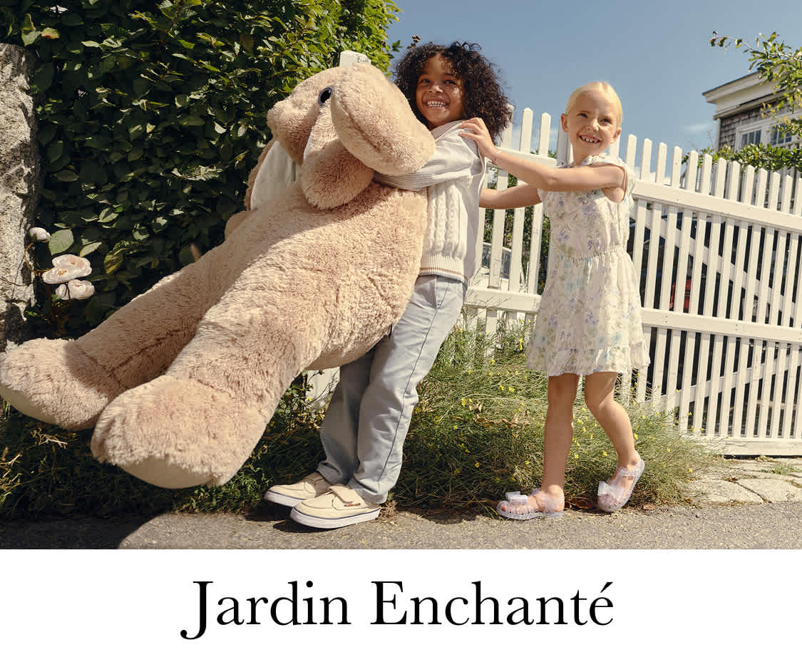 A vintage light blue Volkswagen Beetle parked in a lush, verdant garden setting, with two young women dressed in flowing, bohemian-style outfits standing next to the car.