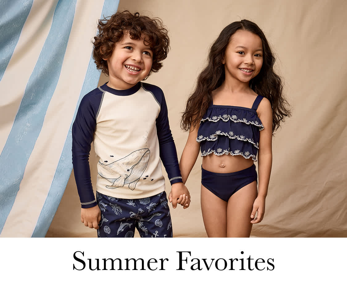 Two young girls with curly hair, one wearing a navy blue shirt and the other a pink top, standing in a room with potted plants in the background.