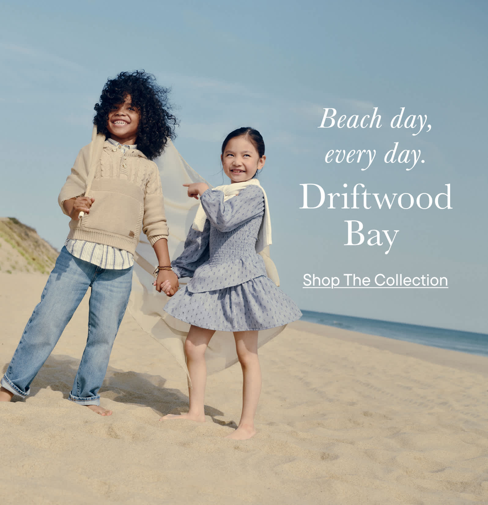 Two young girls, one with curly hair and the other with a dress, are standing on a sandy beach with the ocean and a clear sky in the background.