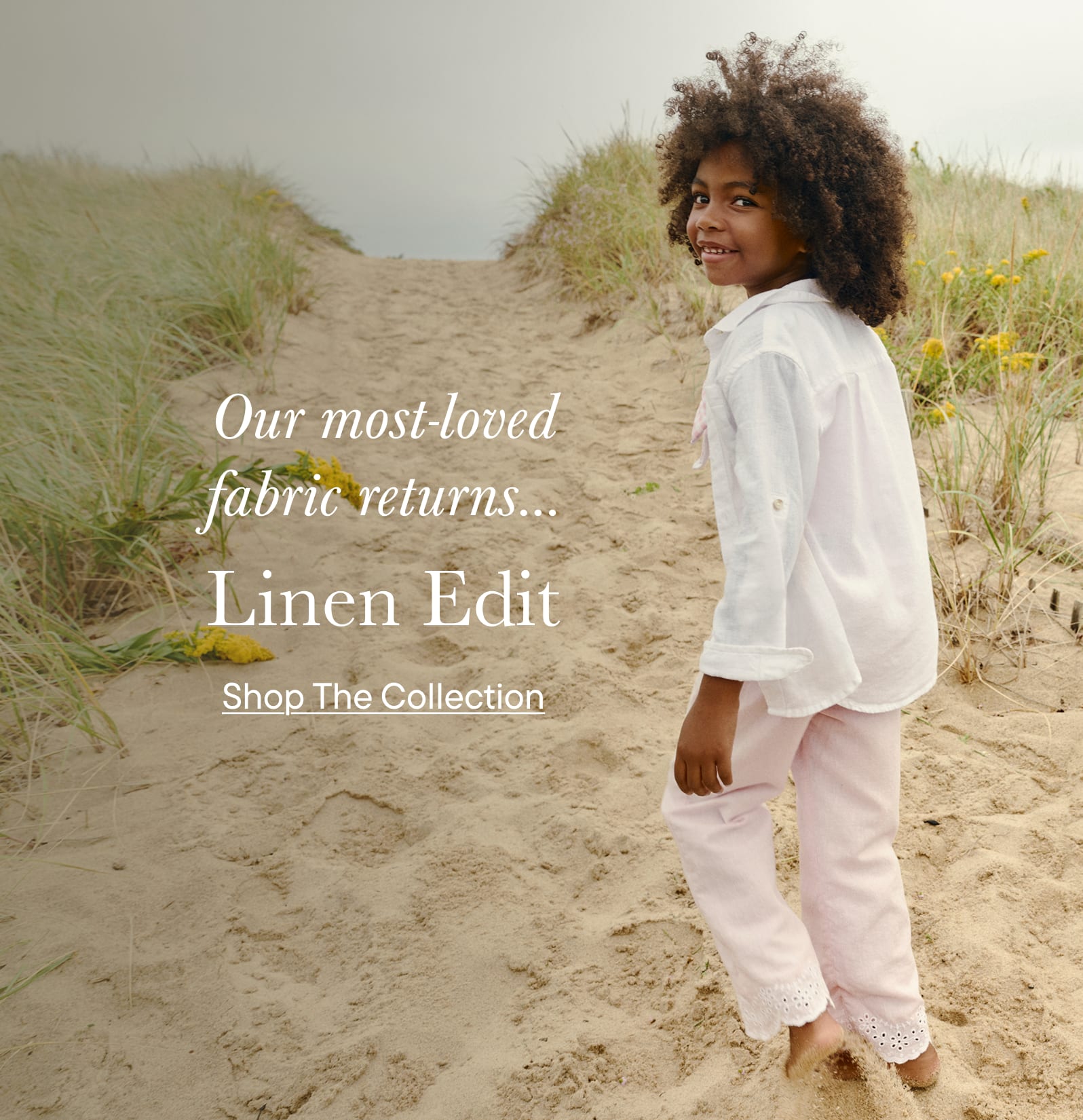A smiling young woman with curly hair stands on a sandy path surrounded by grassy dunes in a coastal setting.