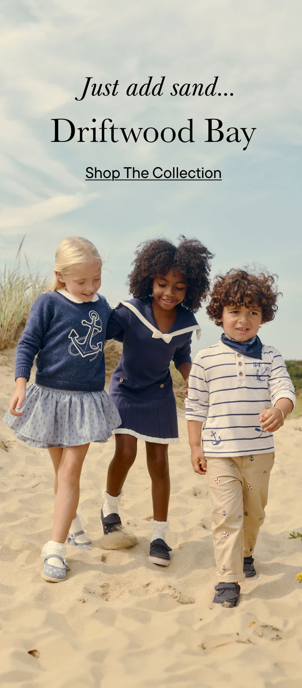 Three young children, a girl and two boys, walking together on a sandy beach with driftwood in the background.