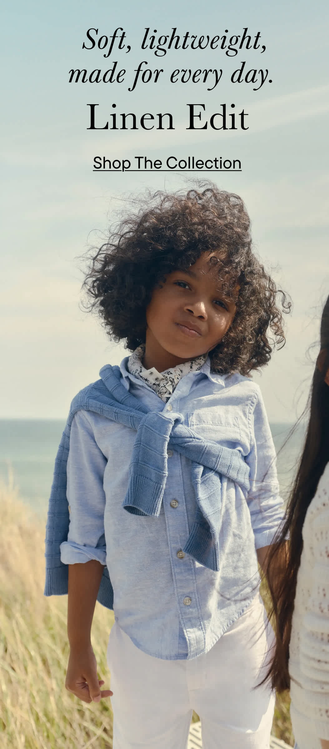 A young woman with curly hair stands in a grassy field, wearing a denim jacket and smiling at the camera.