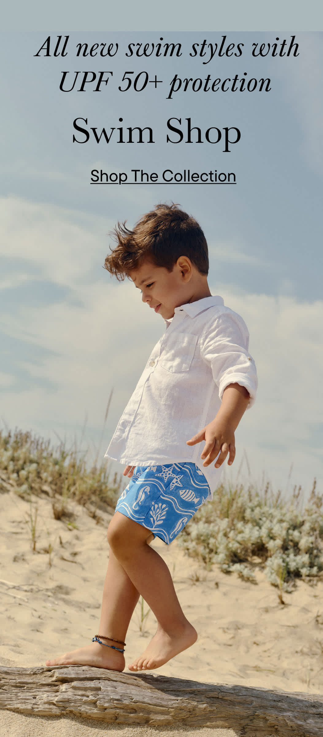 A young boy in white shirt and blue patterned swim trunks running on a sandy beach with a cloudy sky in the background.