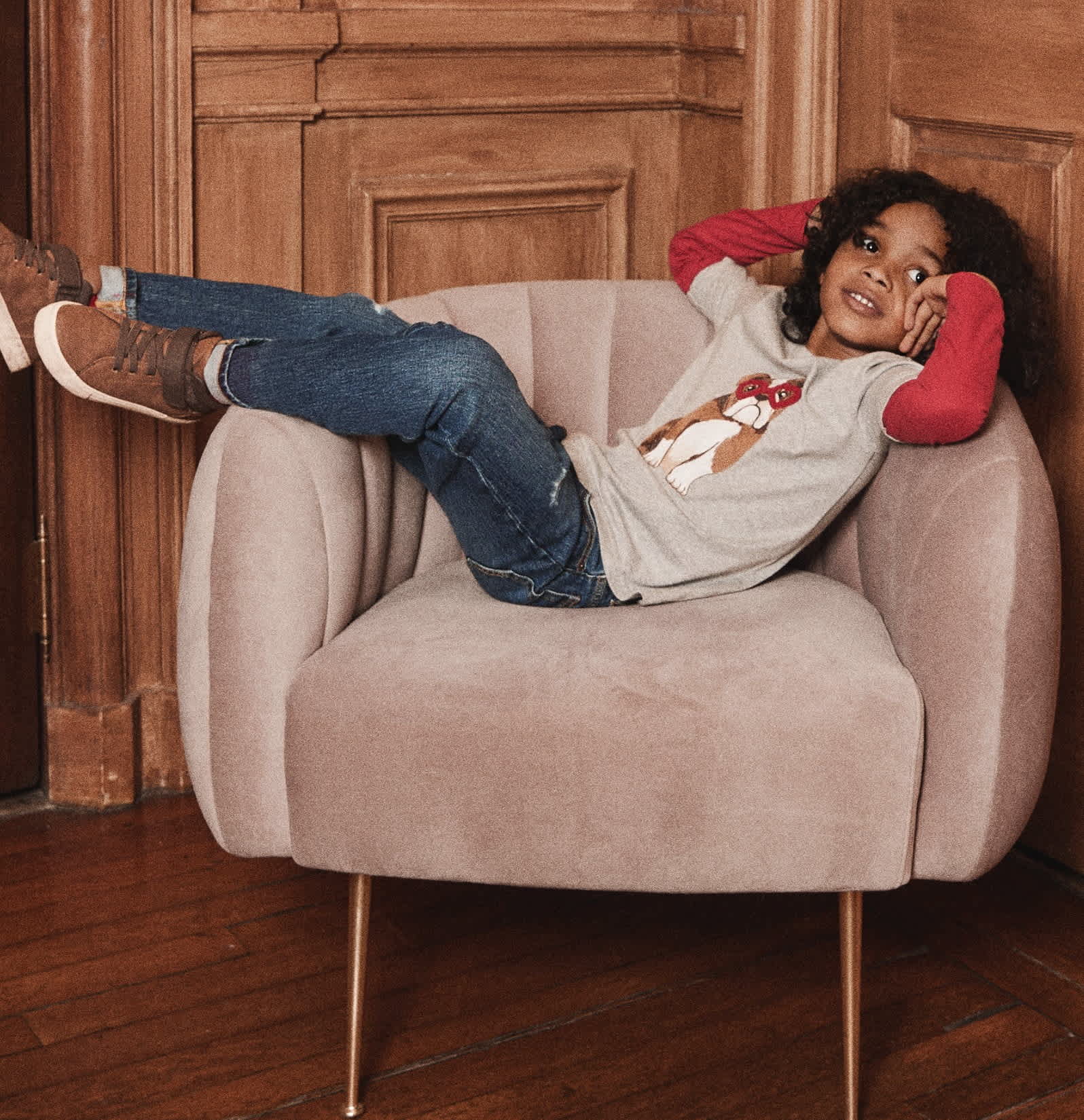 Une jeune femme aux cheveux bouclés est confortablement installée dans un fauteuil beige moelleux, dans une pièce aux murs lambrissés.