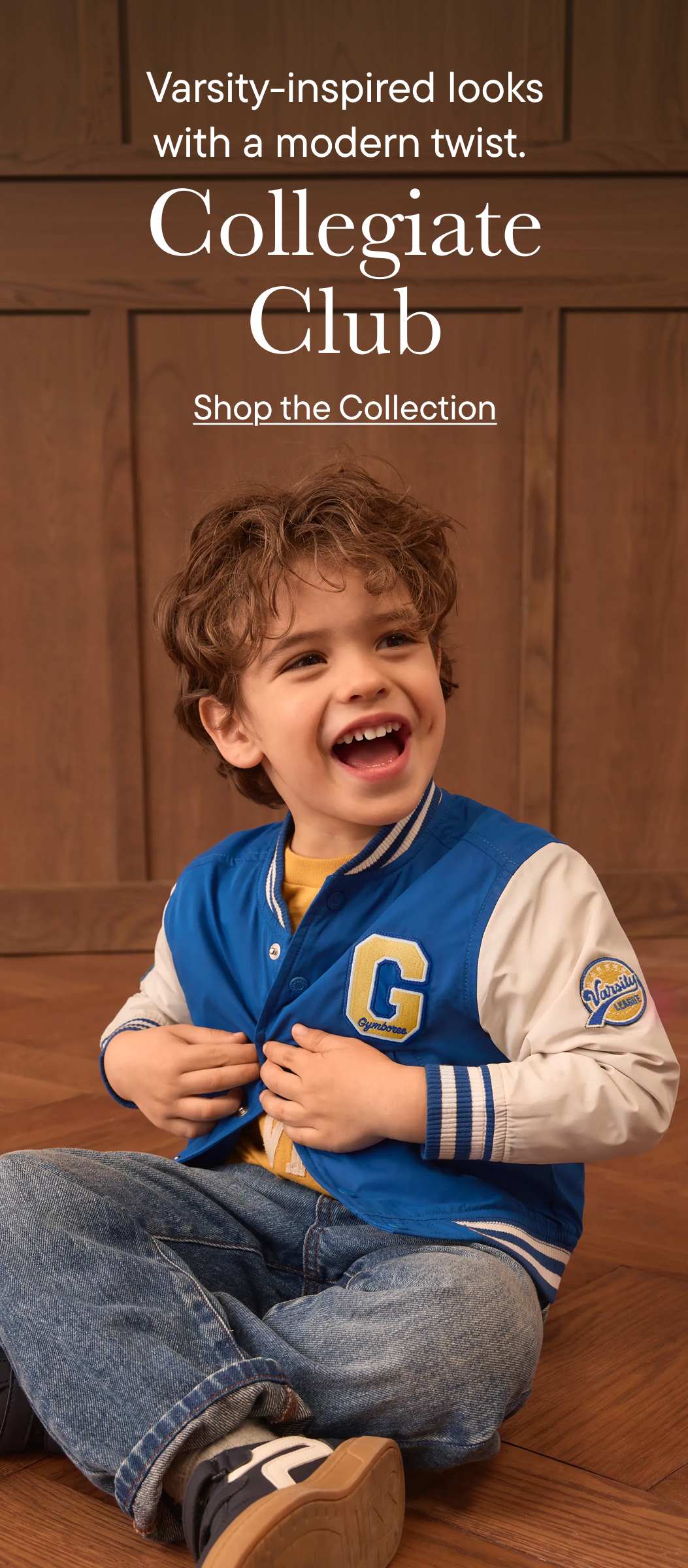 A young boy in a blue and white varsity jacket and jeans sits on a wooden floor in front of a paneled wooden wall.
