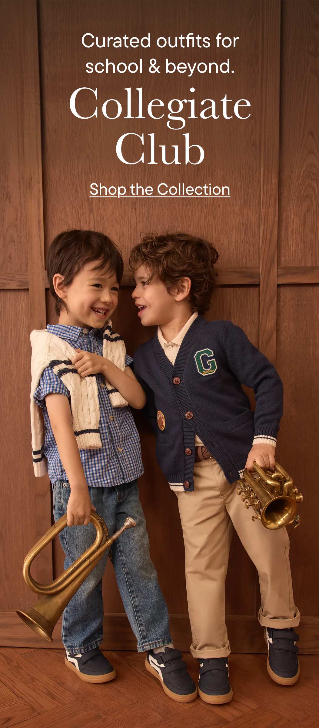 Two young boys in collegiate-style outfits stand holding brass instruments against a warm wooden background.