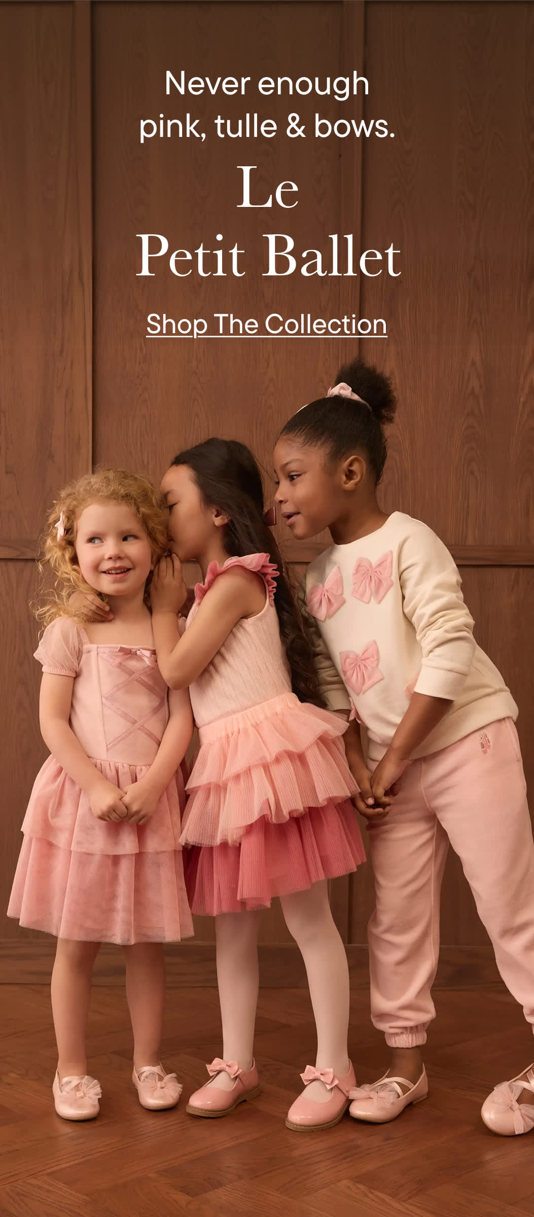 Three young girls in pink dresses and outfits whisper secrets in front of a wooden paneled background.