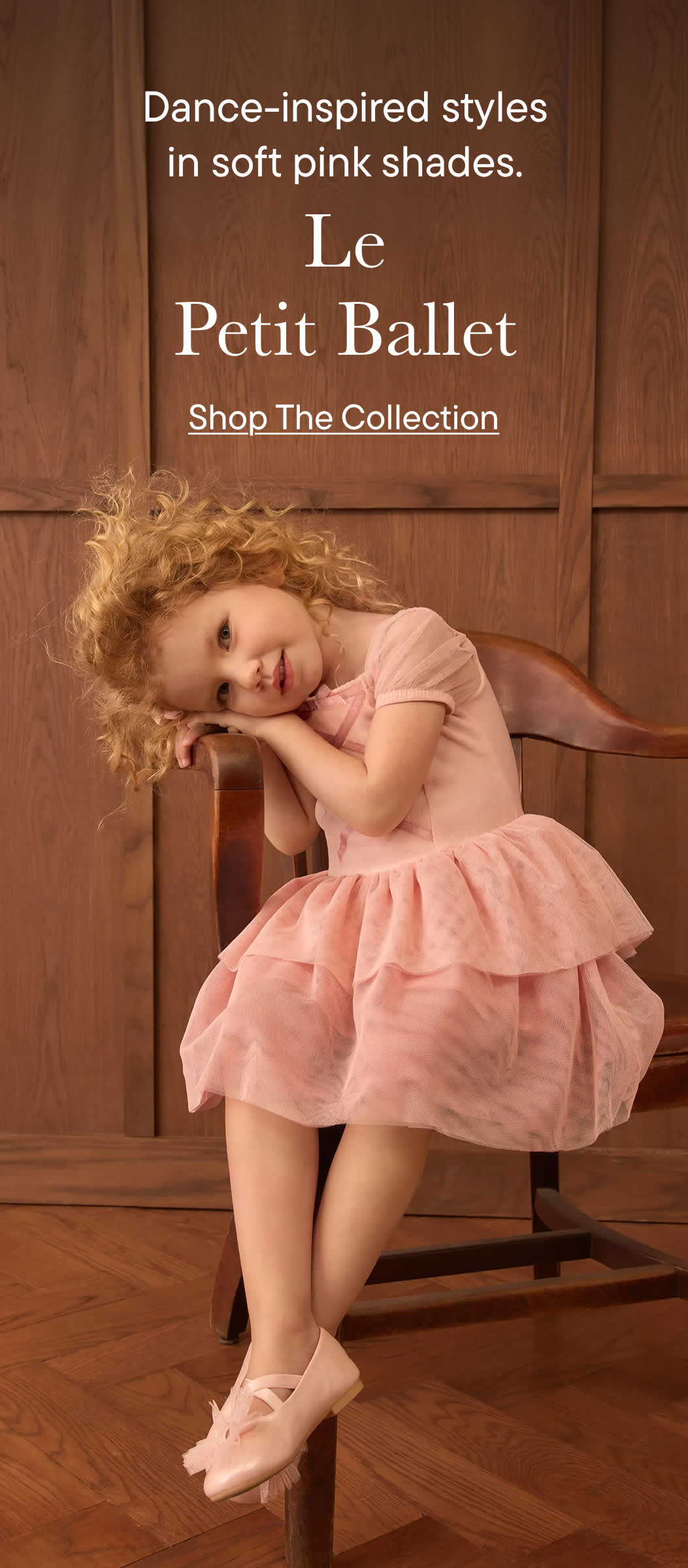 Una niña con un vestido de ballet rosa está sentada en una silla de madera frente a un fondo revestido de paneles de madera.