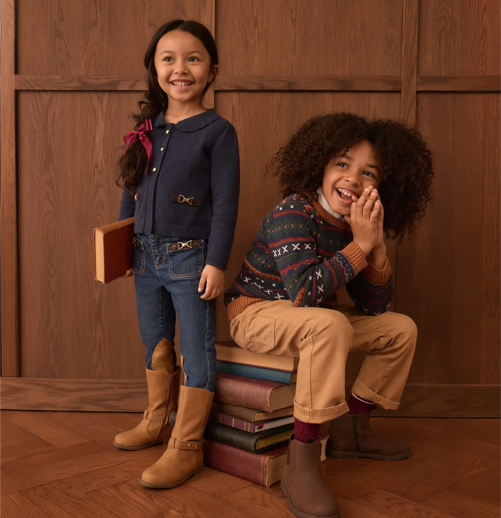 Two smiling children, one standing with a book and the other sitting on a stack of books, are posed against a warm, wood-paneled background.