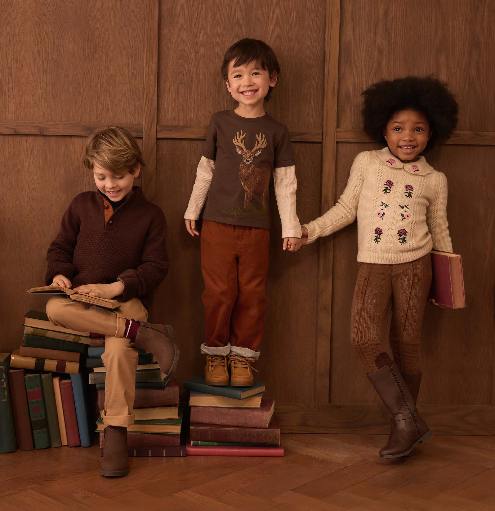 Three children in autumnal clothing are posed with books in front of a wooden paneled wall.