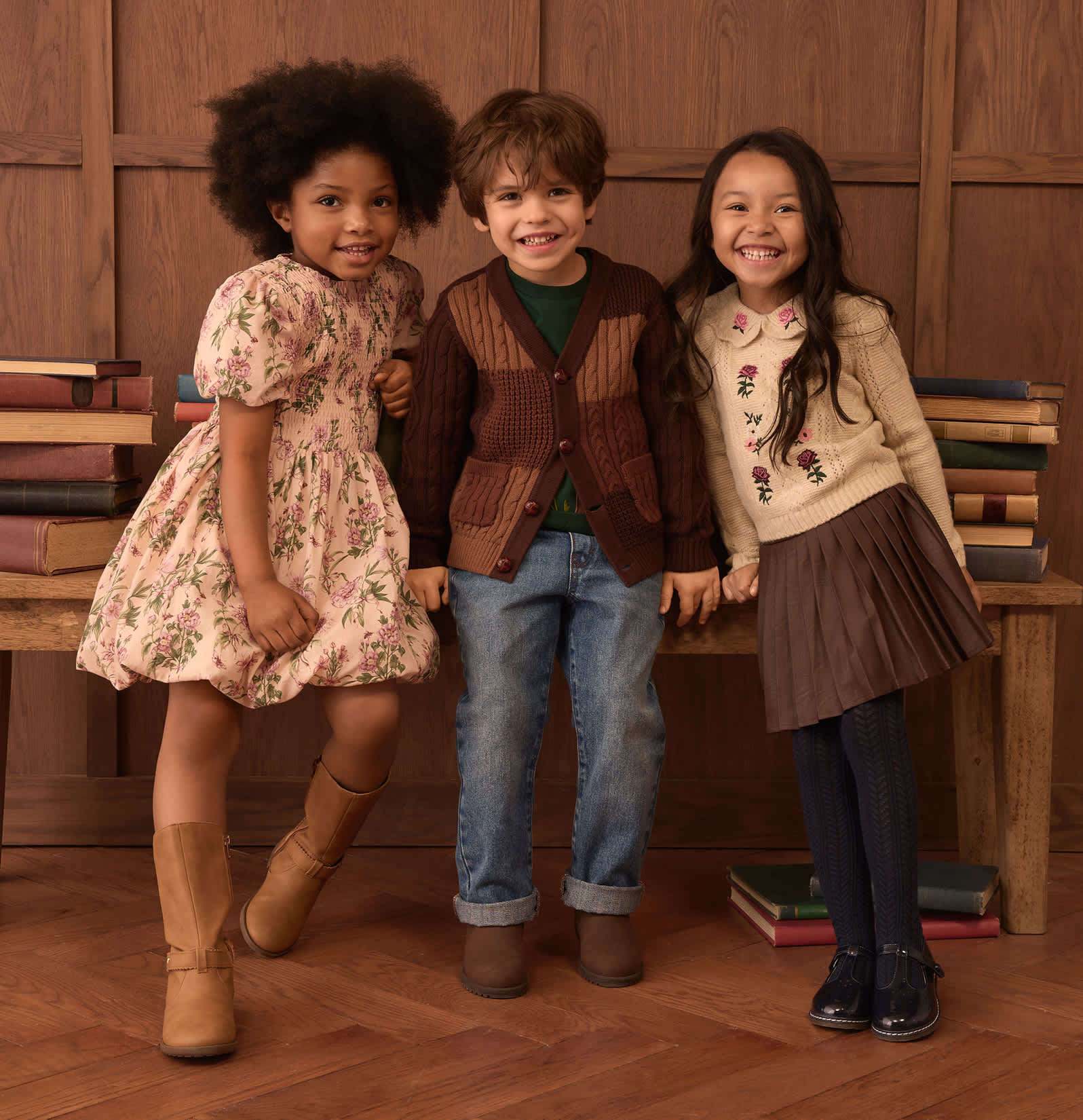 Three smiling children, two girls and a boy, stand in front of a wooden bench laden with books, with a paneled wooden wall behind them.