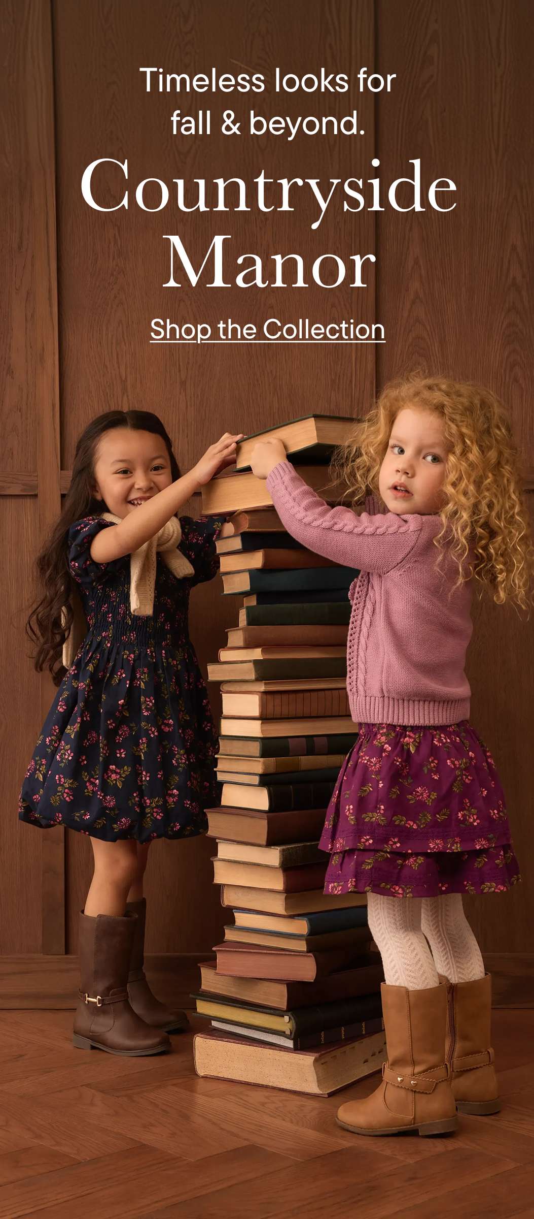 Two young girls reach for a tall stack of books in front of a wood-paneled wall with text.