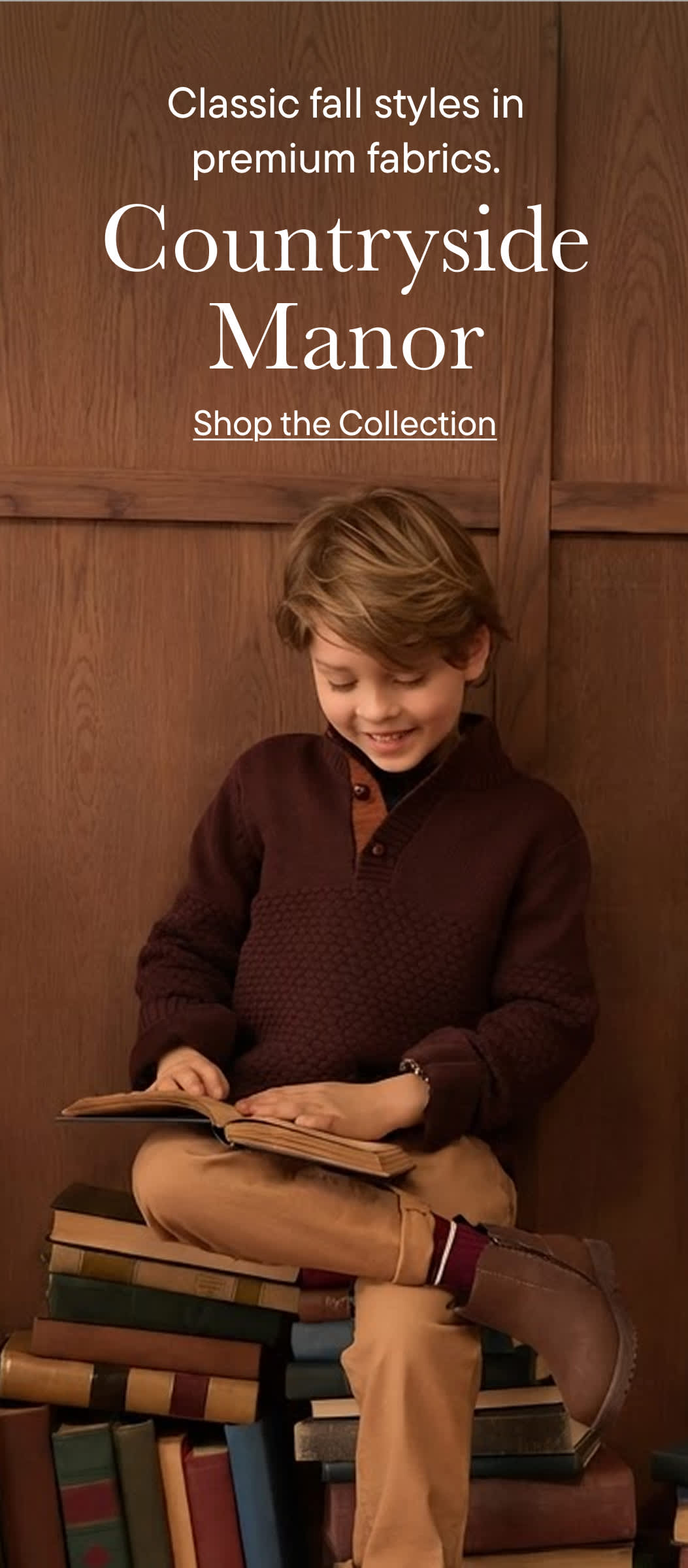 A young boy in fall attire sits on a stack of books, reading, against a wooden background.