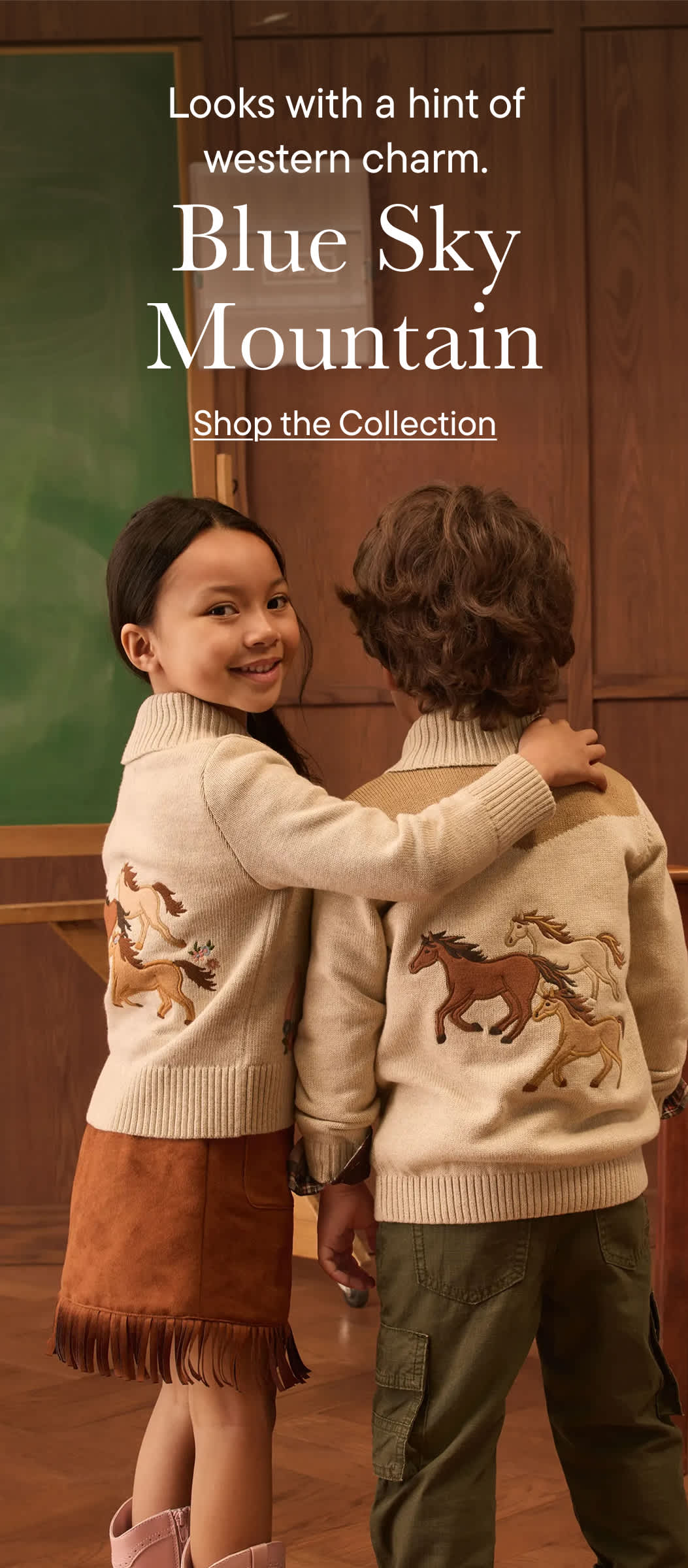 Two children in western-themed sweaters stand in front of a classroom with a chalkboard and wooden paneling.
