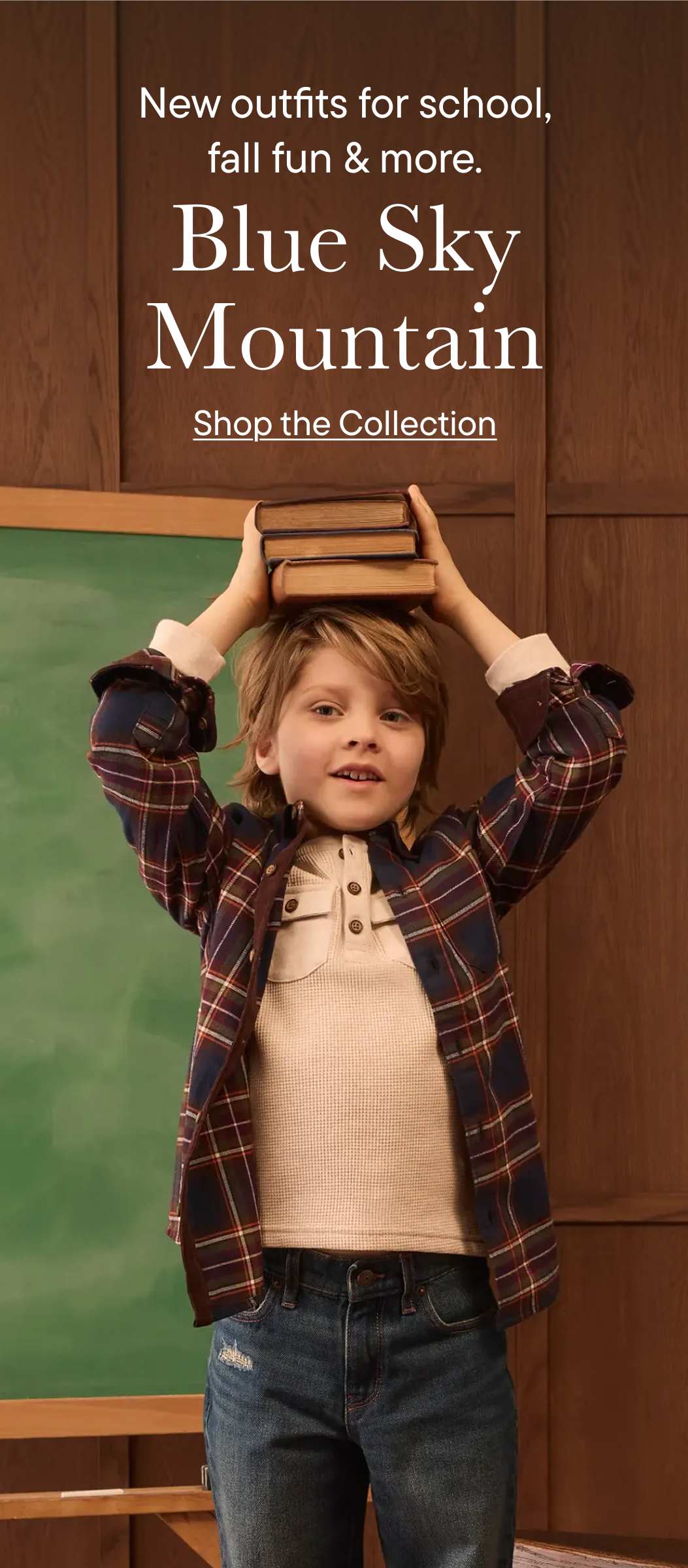 A young boy in a plaid shirt and jeans balances books on his head in front of a classroom blackboard and wood-paneled wall.