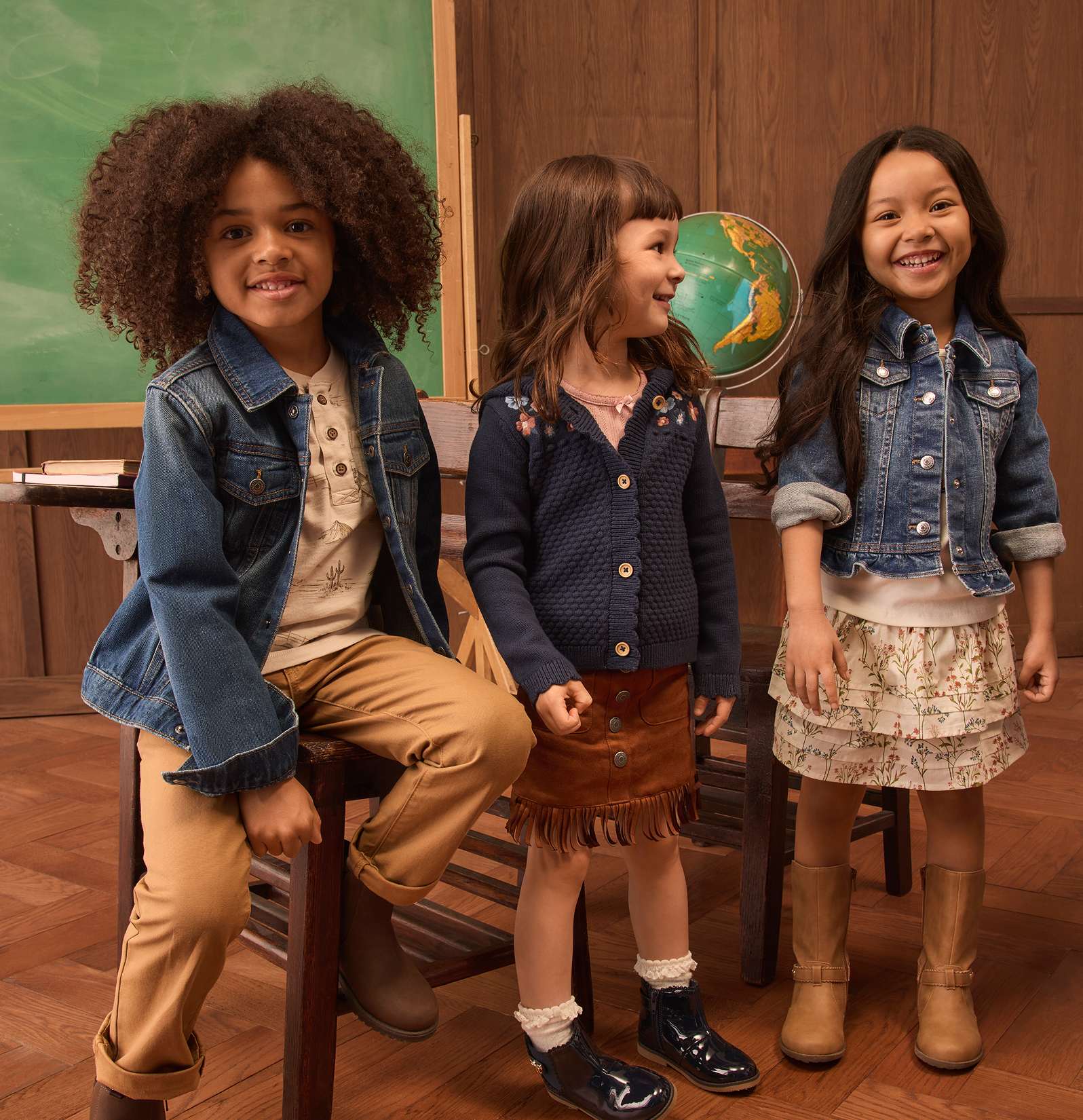 Tres niños sonrientes con ropa informal posan frente a un aula con un globo terráqueo y una pizarra.