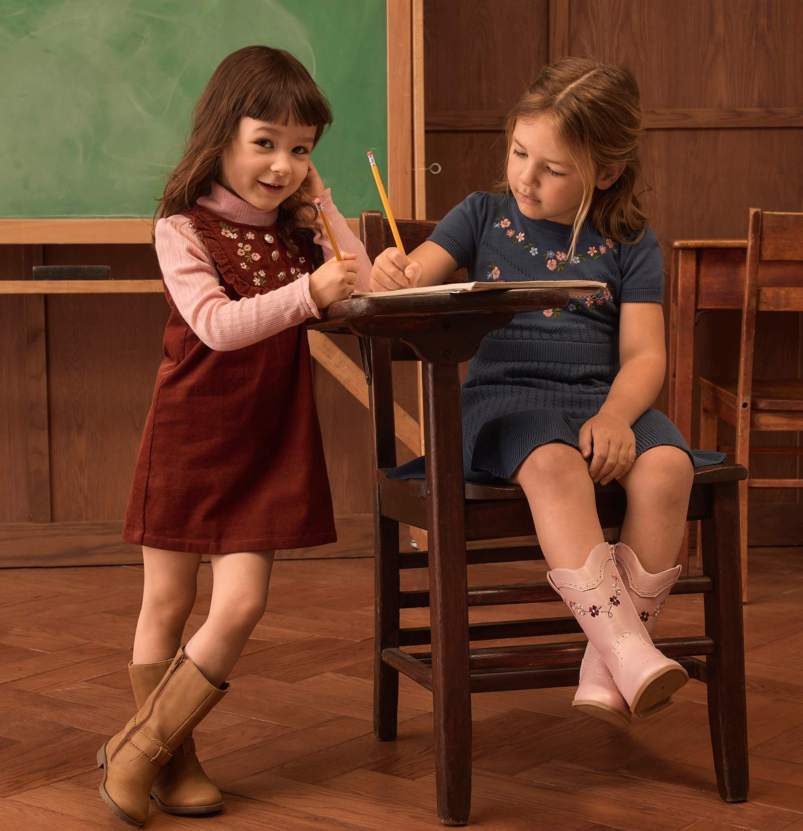 Dos niñas pequeñas, una de pie con un lápiz en la mano y la otra sentada escribiendo en un pupitre, están en un aula con pizarra y muebles de madera.