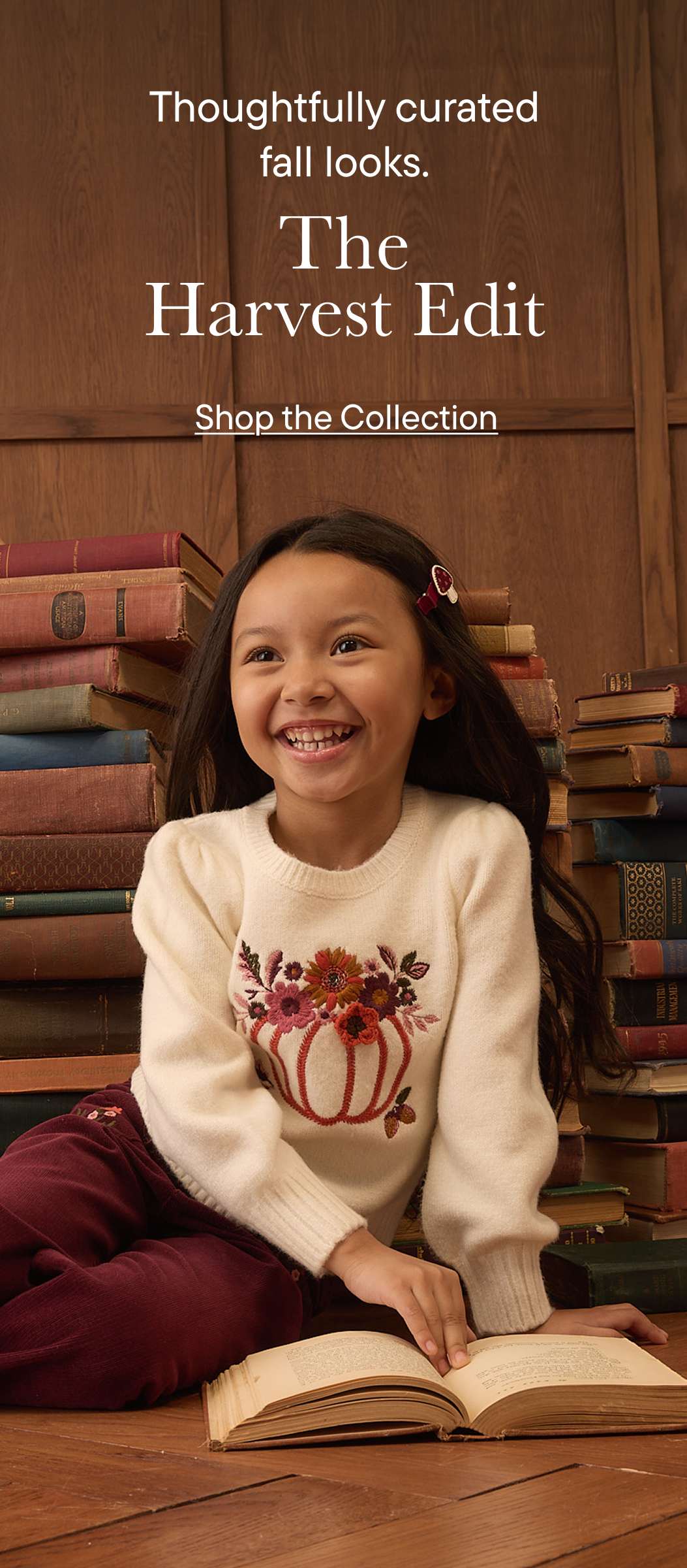 A smiling young girl in a cream sweater with a pumpkin design sits on a wooden floor reading a book, surrounded by stacks of old books against a wooden paneled background.