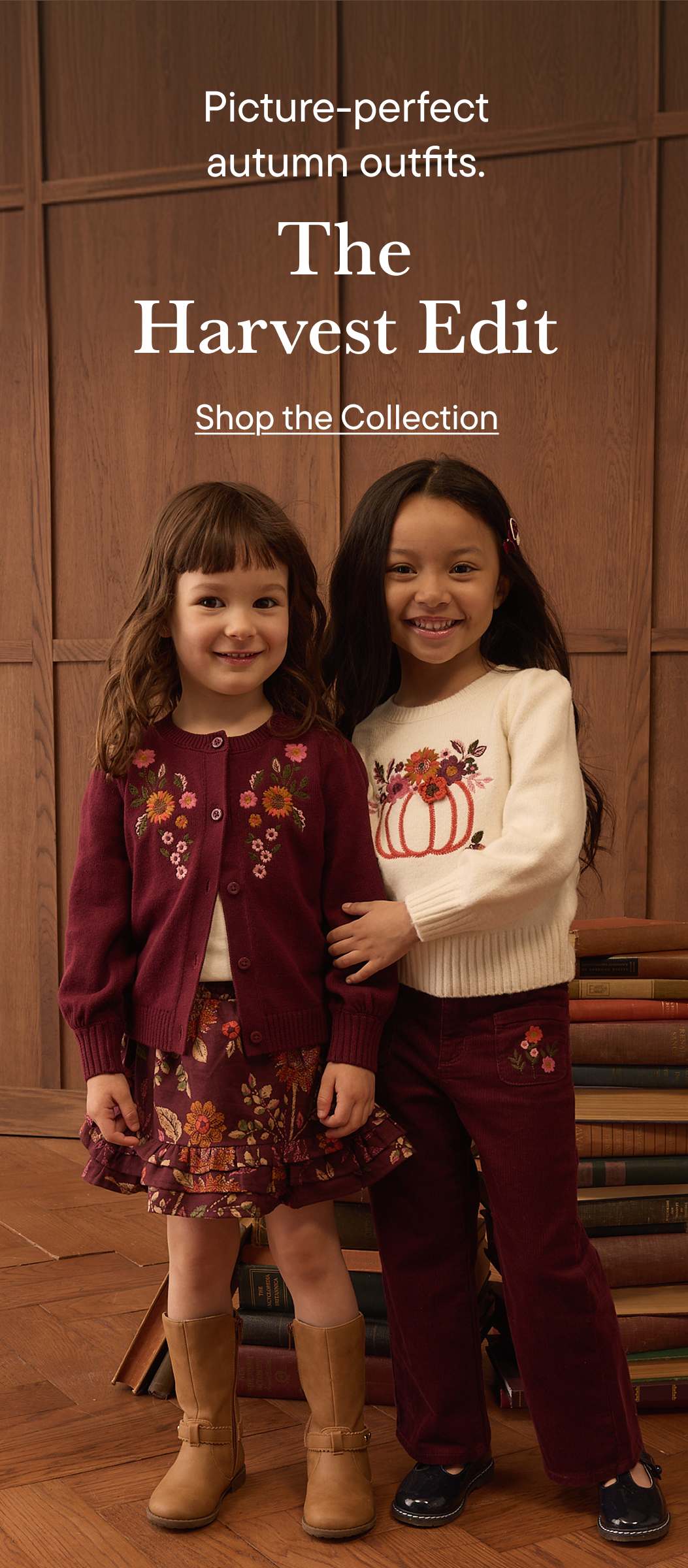 Two smiling young girls in autumn outfits stand in front of a wall of books.