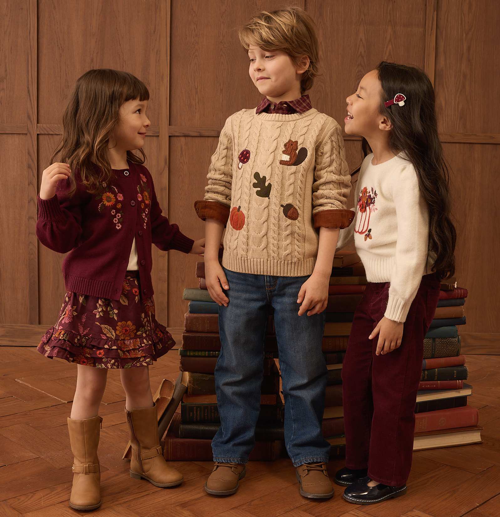 Tres niños vestidos con ropa de otoño están de pie frente a una pila de libros y una pared revestida de madera.