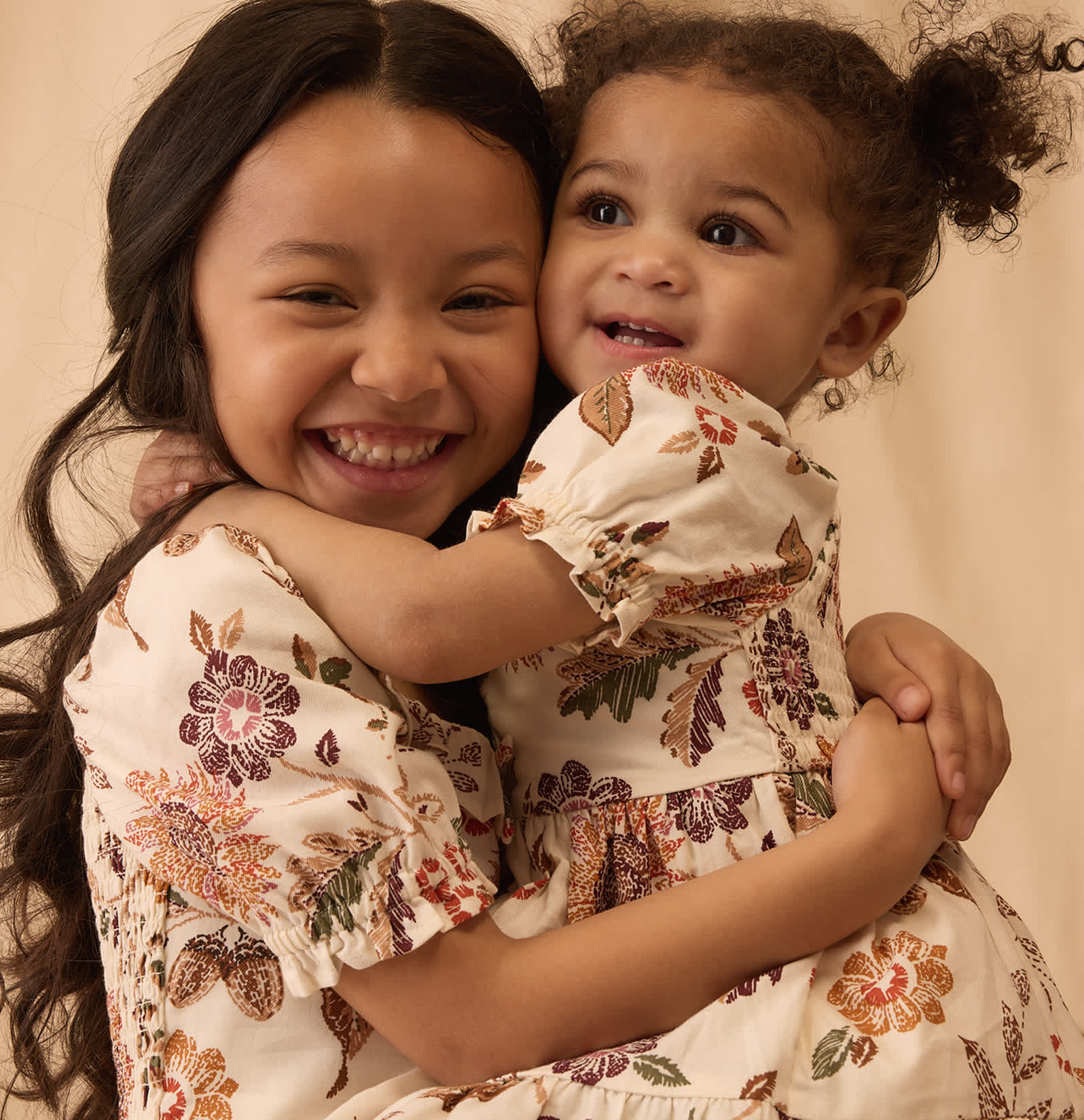 Dos niñas pequeñas con vestidos florales se abrazan fuertemente frente a un fondo liso de color claro.