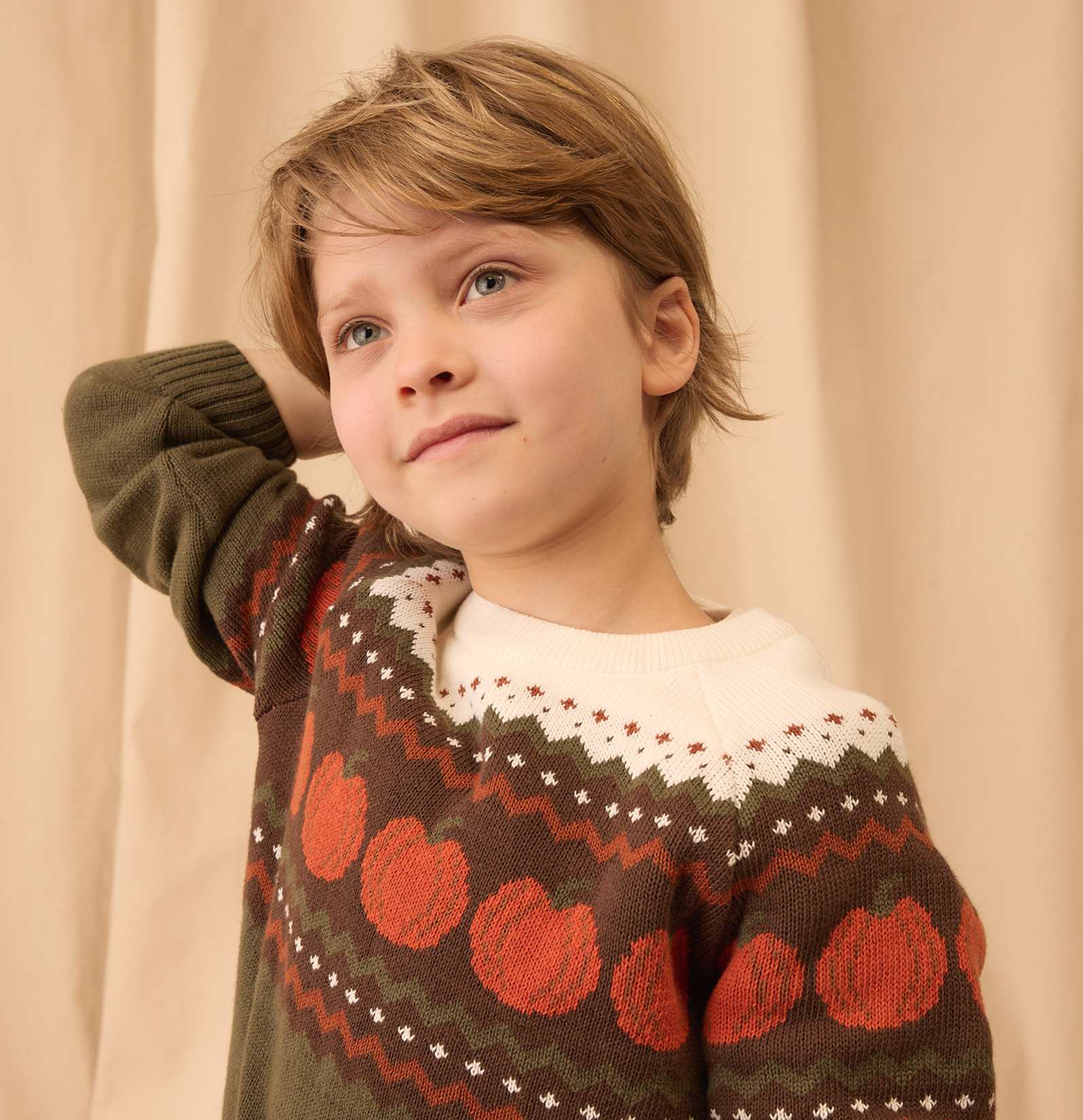 Un niño pequeño, rubio y de ojos azules, que lleva un suéter estampado, mira hacia arriba y hacia un lado, con un fondo suave de color beige.