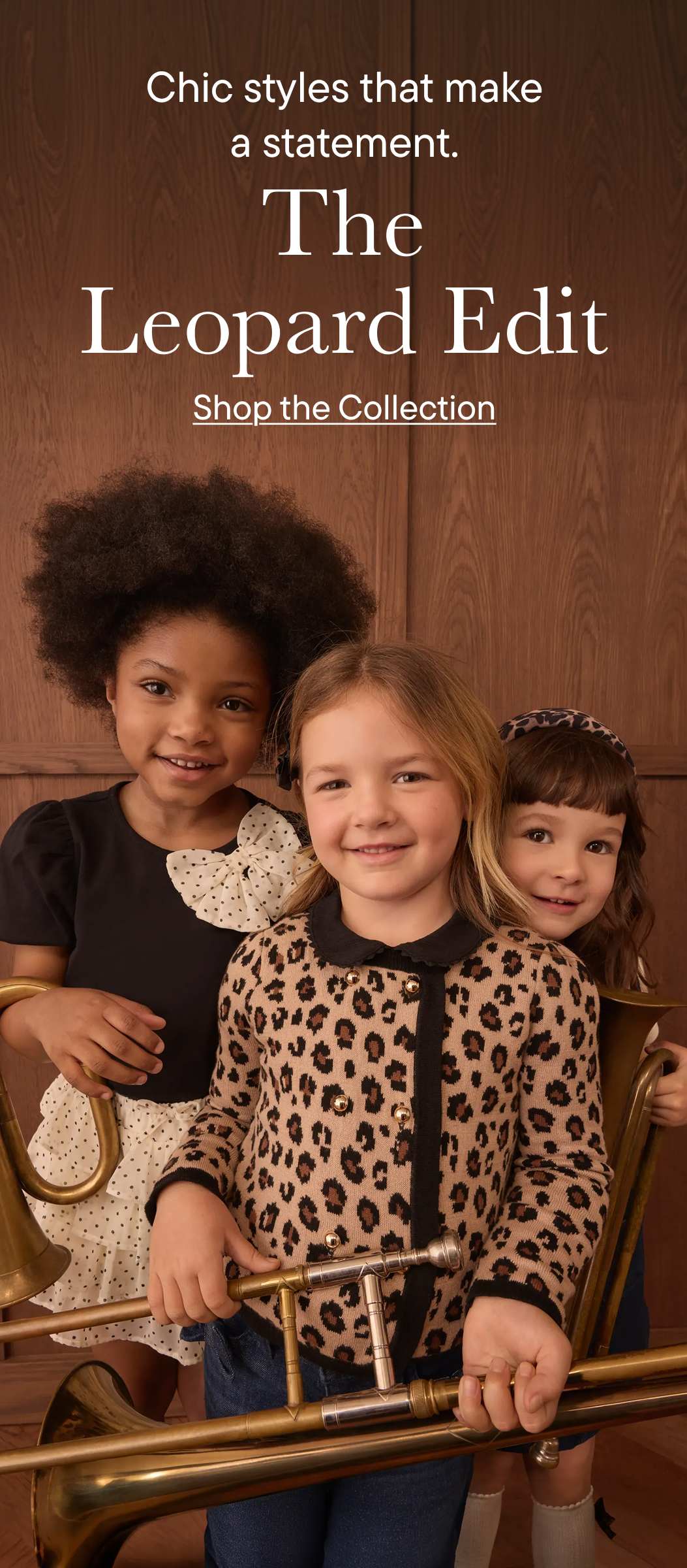 Tres niñas vestidas con ropa de estampado de leopardo sostienen instrumentos de viento-metal frente a un fondo revestido de madera.