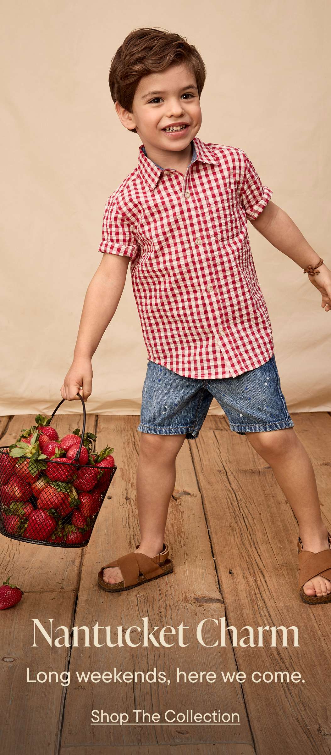 A young boy in a red and white checkered shirt stands on a wooden floor, holding a basket filled with fresh strawberries.