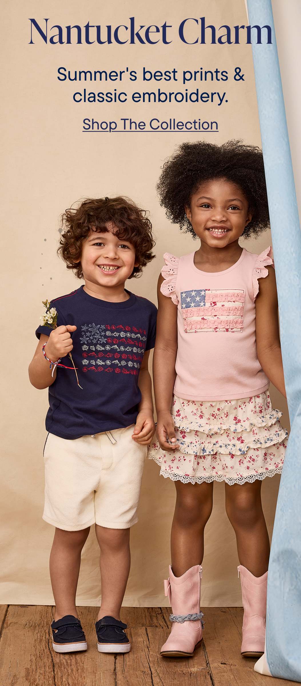 La imagen muestra a dos niños pequeños, un niño y una niña, de pie y sonriendo. Llevan ropa veraniega: el niño una camisa azul marino y la niña un vestido rosa y blanco estampado. El fondo parece ser un anuncio publicitario de una colección de ropa.