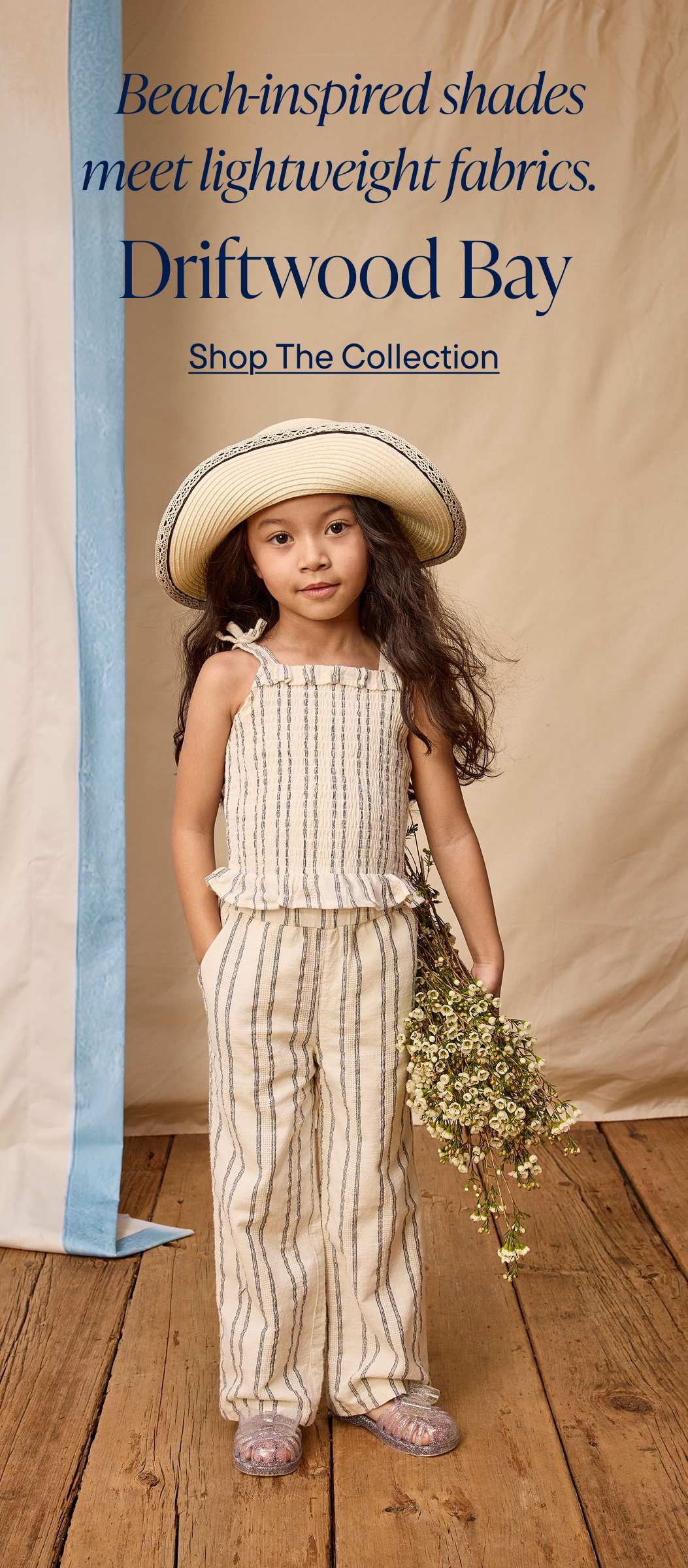 Una jovencita con un sombrero de ala ancha y un conjunto de rayas posa frente a una pancarta que anuncia la colección de ropa de Driftwood Bay.