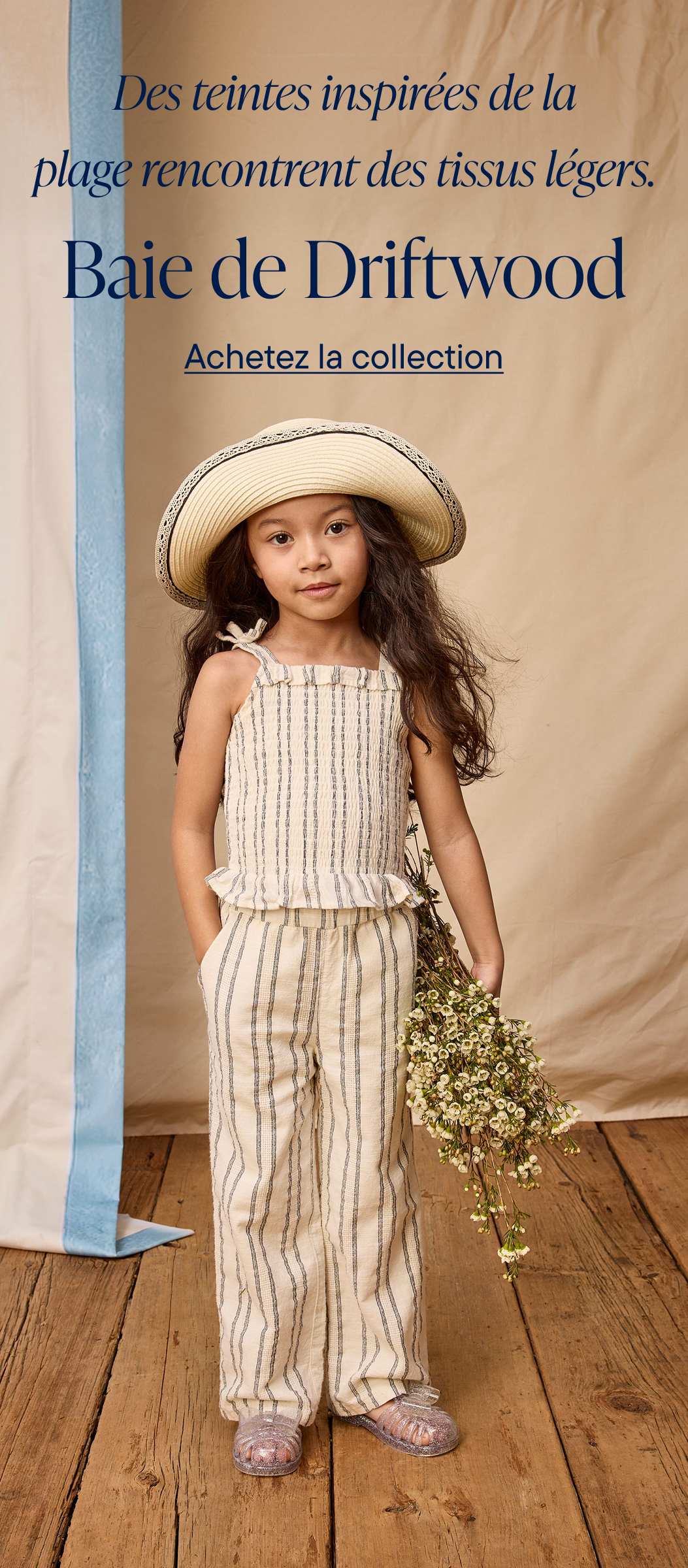 A young girl wearing a wide-brimmed hat and a striped outfit stands in front of a banner advertising Driftwood Bay's clothing collection.