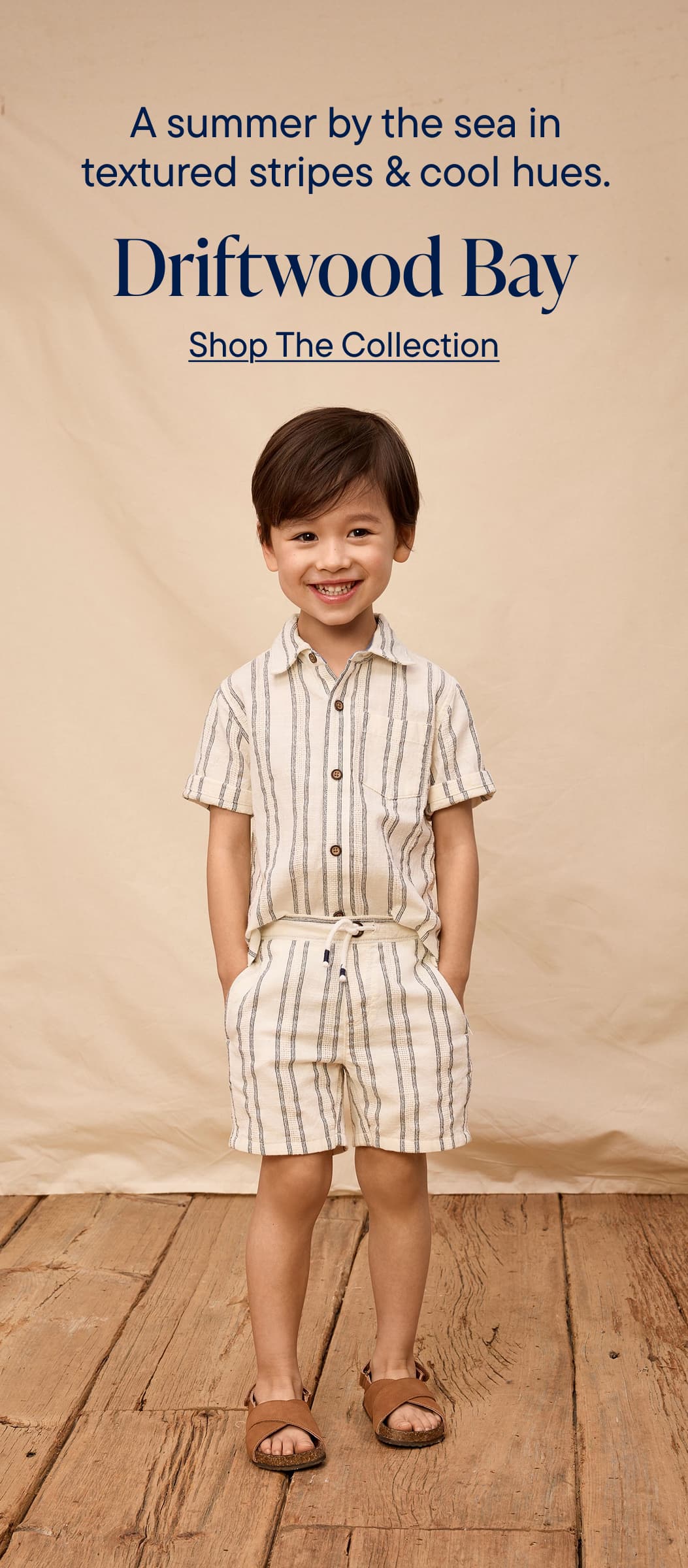 A smiling young person wearing a striped shirt, green jacket, and khaki pants stands on a sandy beach with driftwood in the background, promoting a nautical-inspired clothing collection.