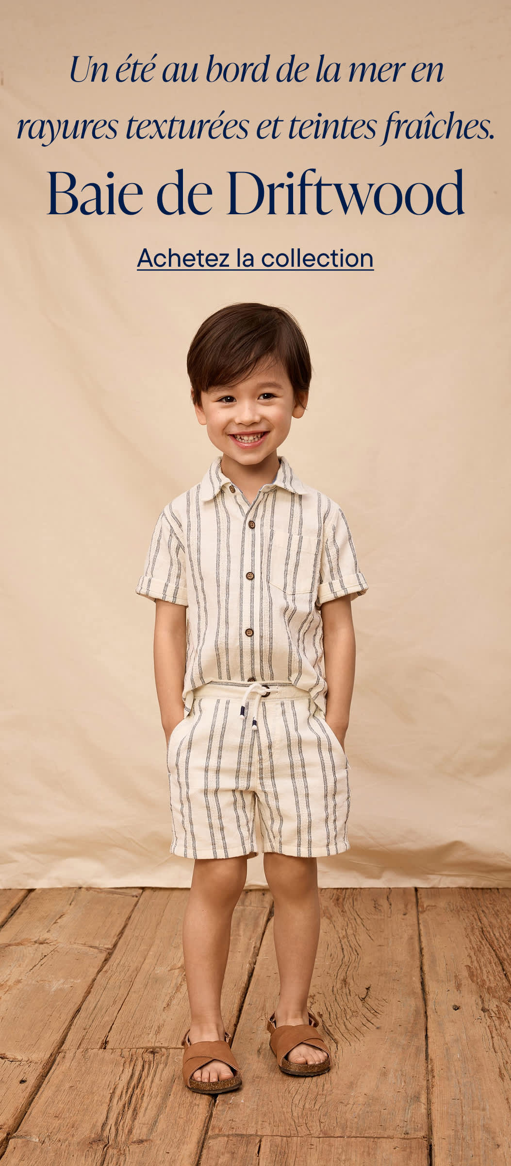 A smiling young person wearing a striped shirt, green jacket, and khaki pants stands on a sandy beach with driftwood in the background, promoting a nautical-inspired clothing collection.