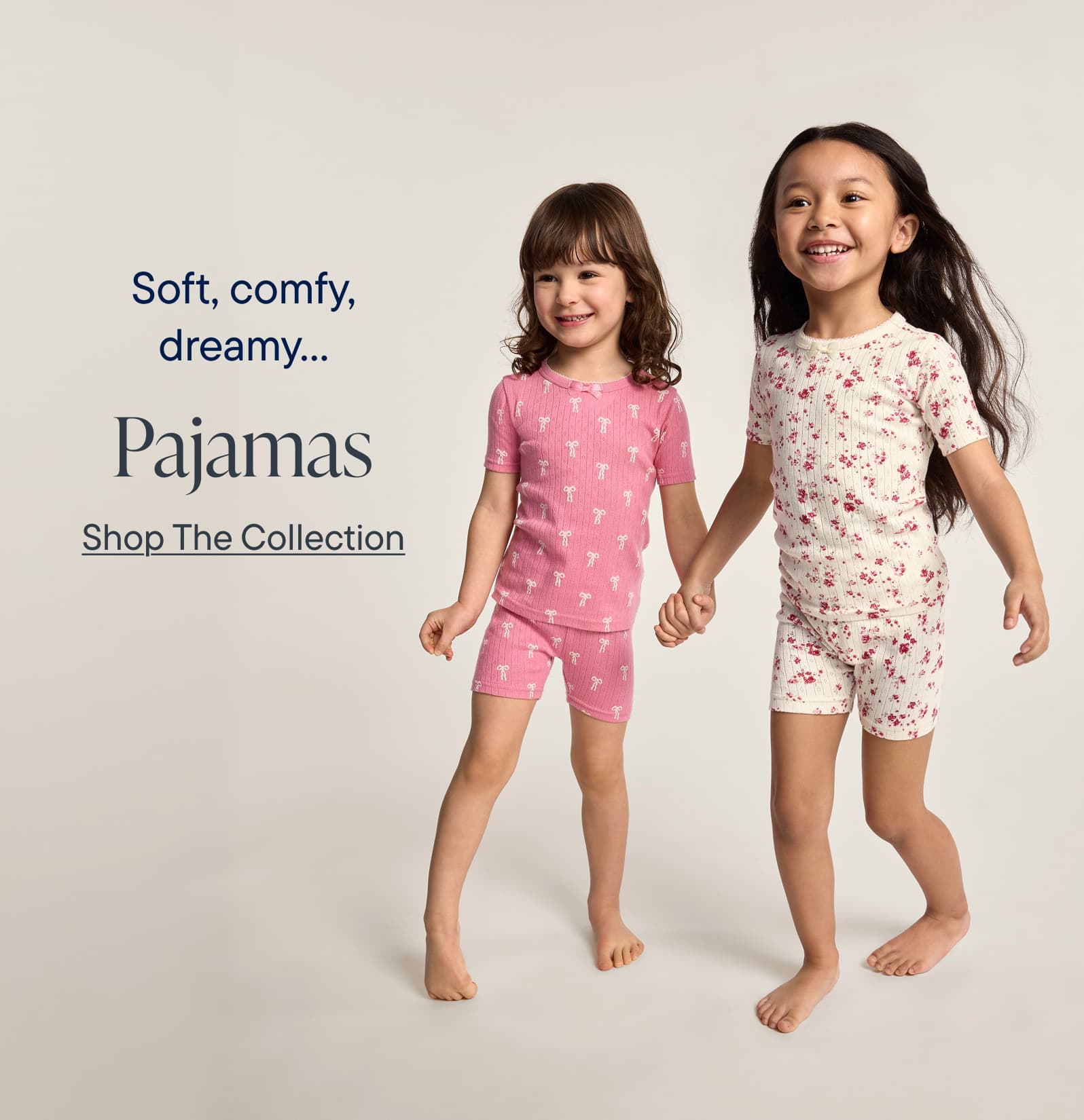 Soft, comfy, dreamy... Pajamas. The image showcases two young girls wearing matching pajama sets, one in a pink polka dot pattern and the other in a floral print. They are standing against a plain, light-colored background, creating a simple and inviting visual.