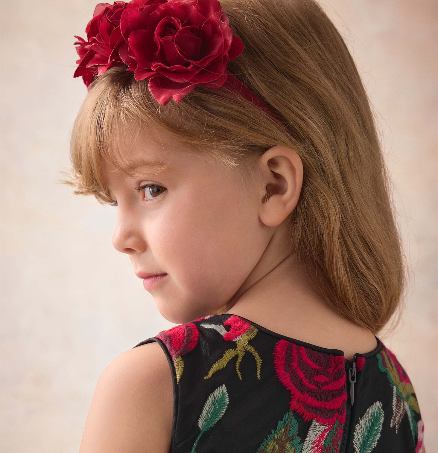 A young girl with a red floral headband and a matching rose-patterned dress is posed against a soft, neutral-toned background.