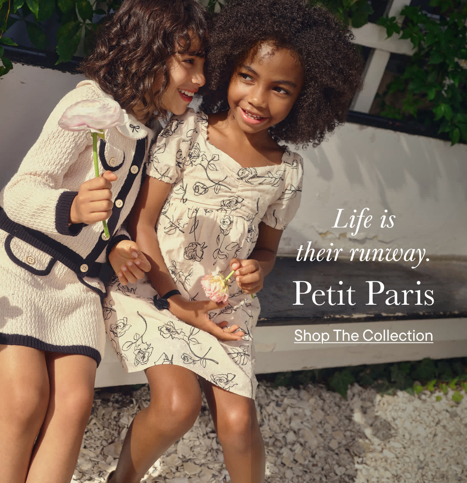 The image shows two young women, one with curly hair and the other with straight hair, standing together and smiling. They appear to be in a natural setting, with greenery visible in the background.