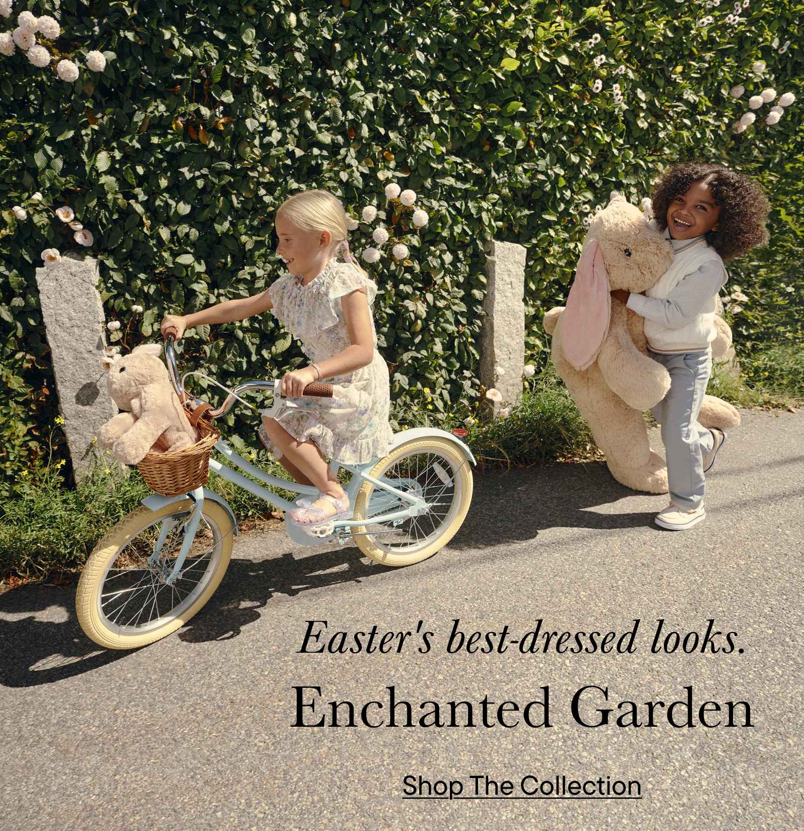 The image depicts two young girls, one riding a bicycle and the other standing beside her, in a lush, verdant garden setting with blooming flowers and greenery in the background. The text overlay promotes an "Enchanted Garden" collection.