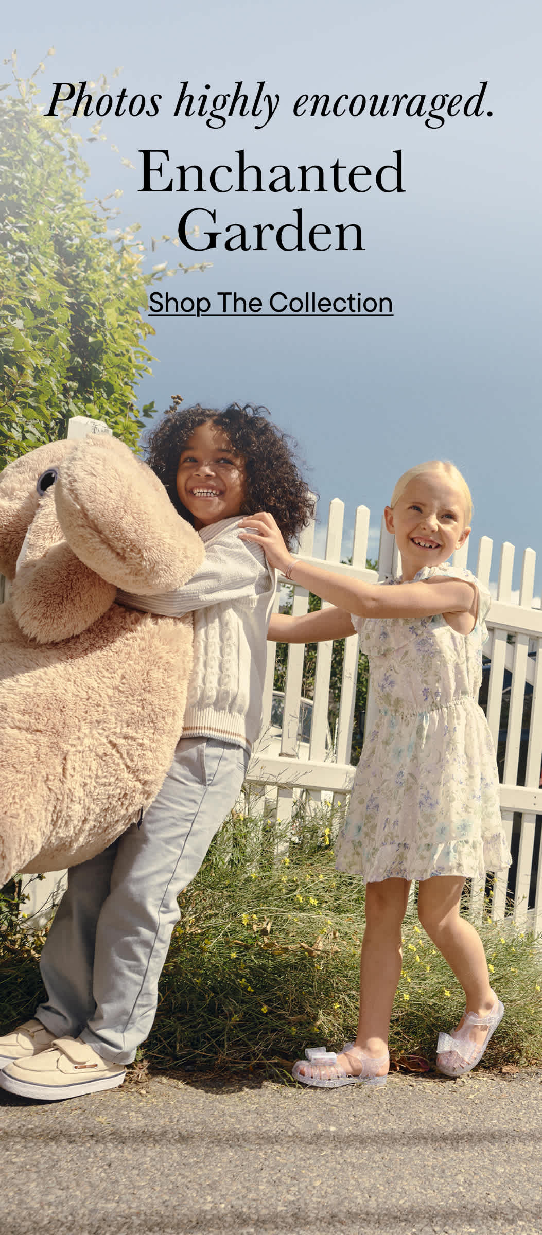 La imagen muestra a dos niños pequeños, una niña con cabello rizado y un niño, jugando juntos en un jardín con una valla blanca al fondo.