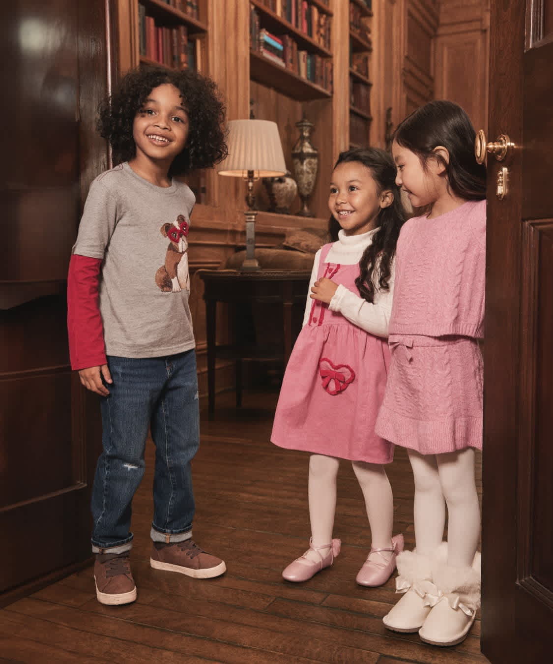 Three young girls, dressed in casual and formal attire, stand together in a cozy, wood-paneled room filled with bookshelves.
