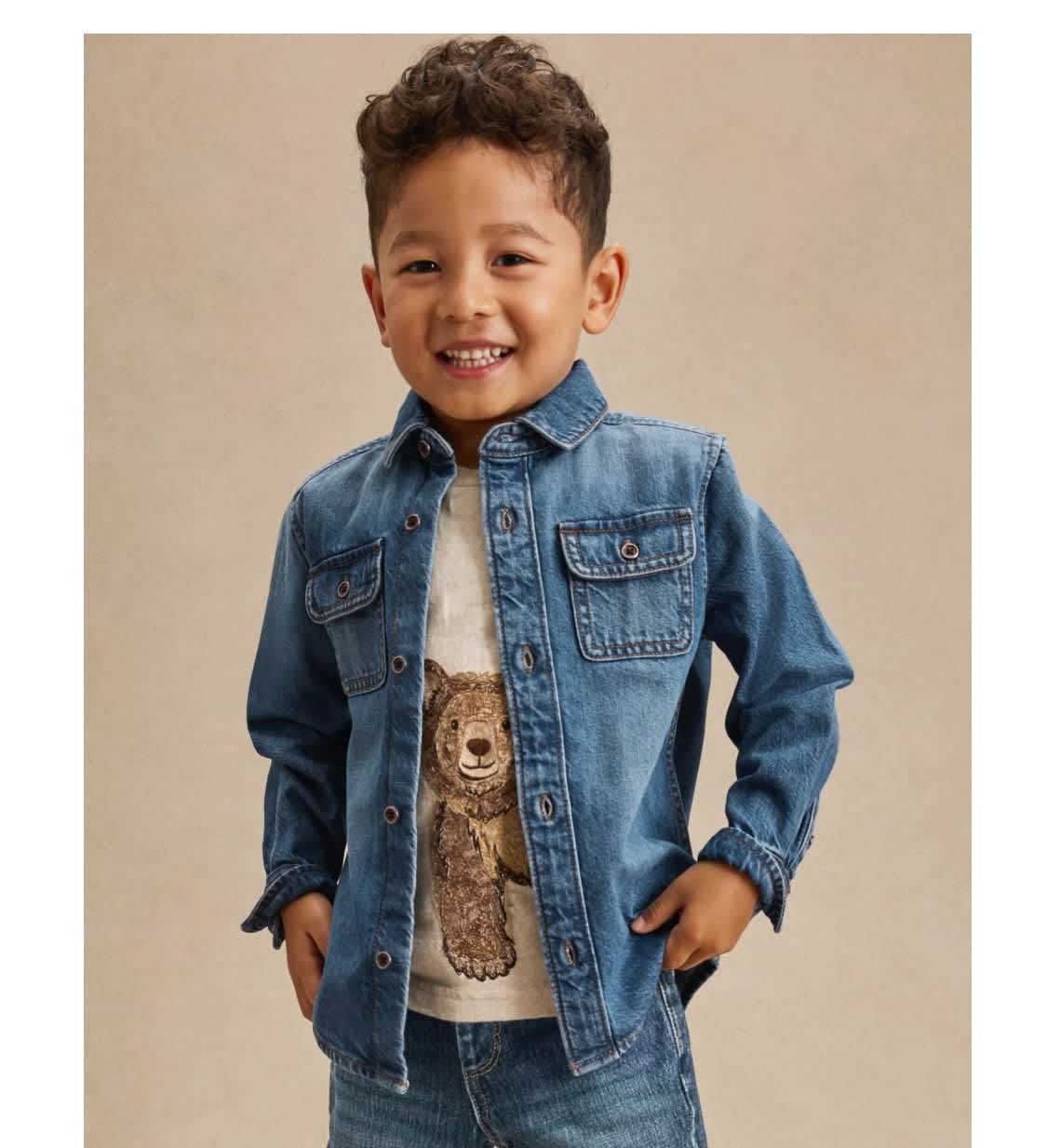 A young boy with curly hair is smiling while wearing a denim jacket and a shirt with a teddy bear design.