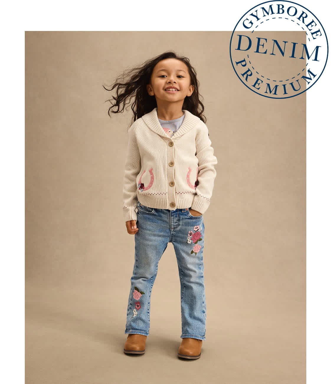 A young girl wearing a white cardigan and embroidered jeans stands against a plain beige background, her hair blowing gently.