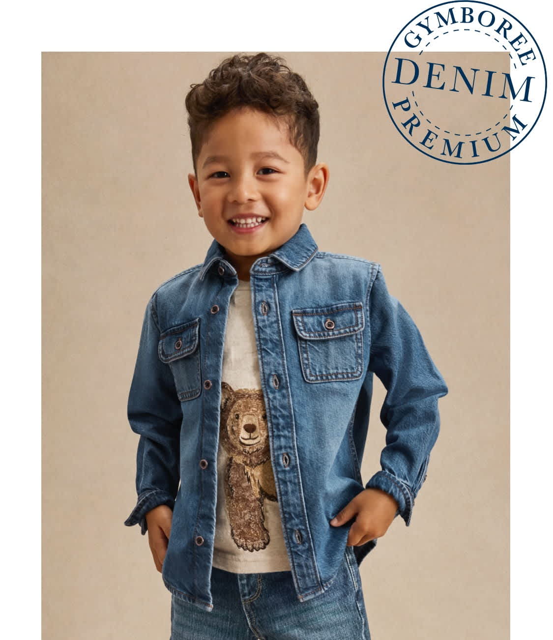 A young boy with curly hair is smiling while wearing a denim shirt and holding a stuffed bear in the foreground, against a plain beige background.