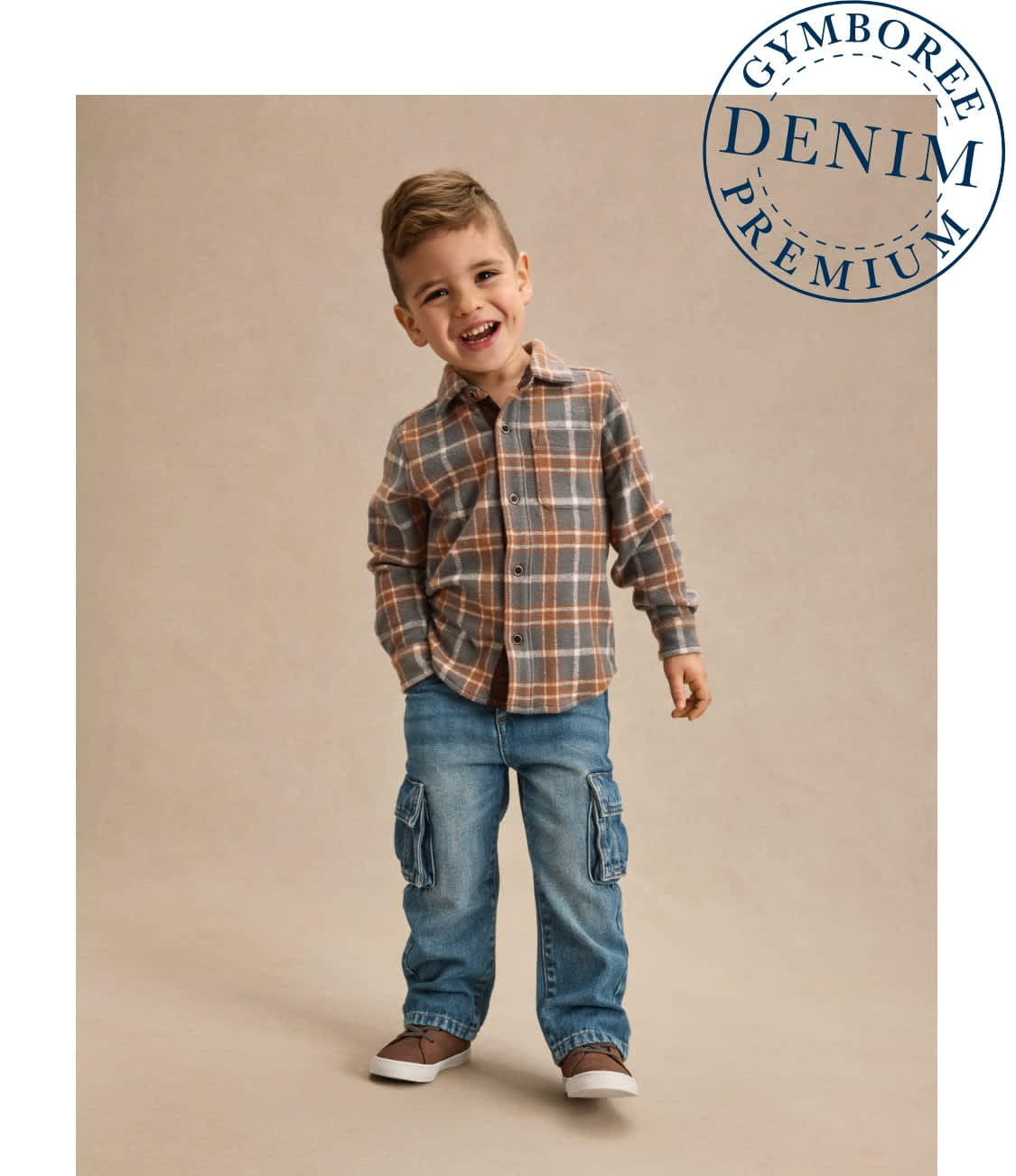 A young boy wearing a plaid shirt and jeans stands against a plain beige background, smiling brightly.