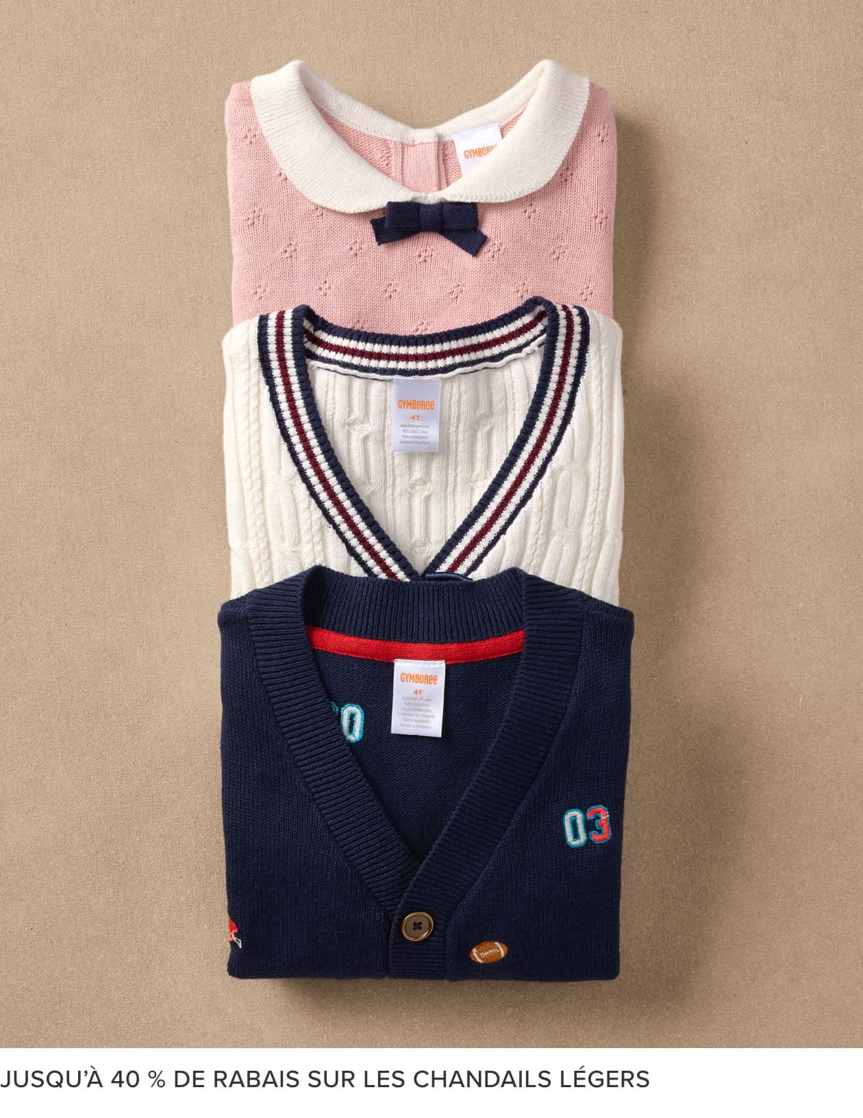 L'image montre une pile de pulls pour enfants soigneusement pliés, de couleurs et de motifs variés, dont un pull rose avec un nœud, un pull rayé et un cardigan bleu marine avec un écusson. Les pulls sont présentés sur un fond beige, probablement dans un magasin.